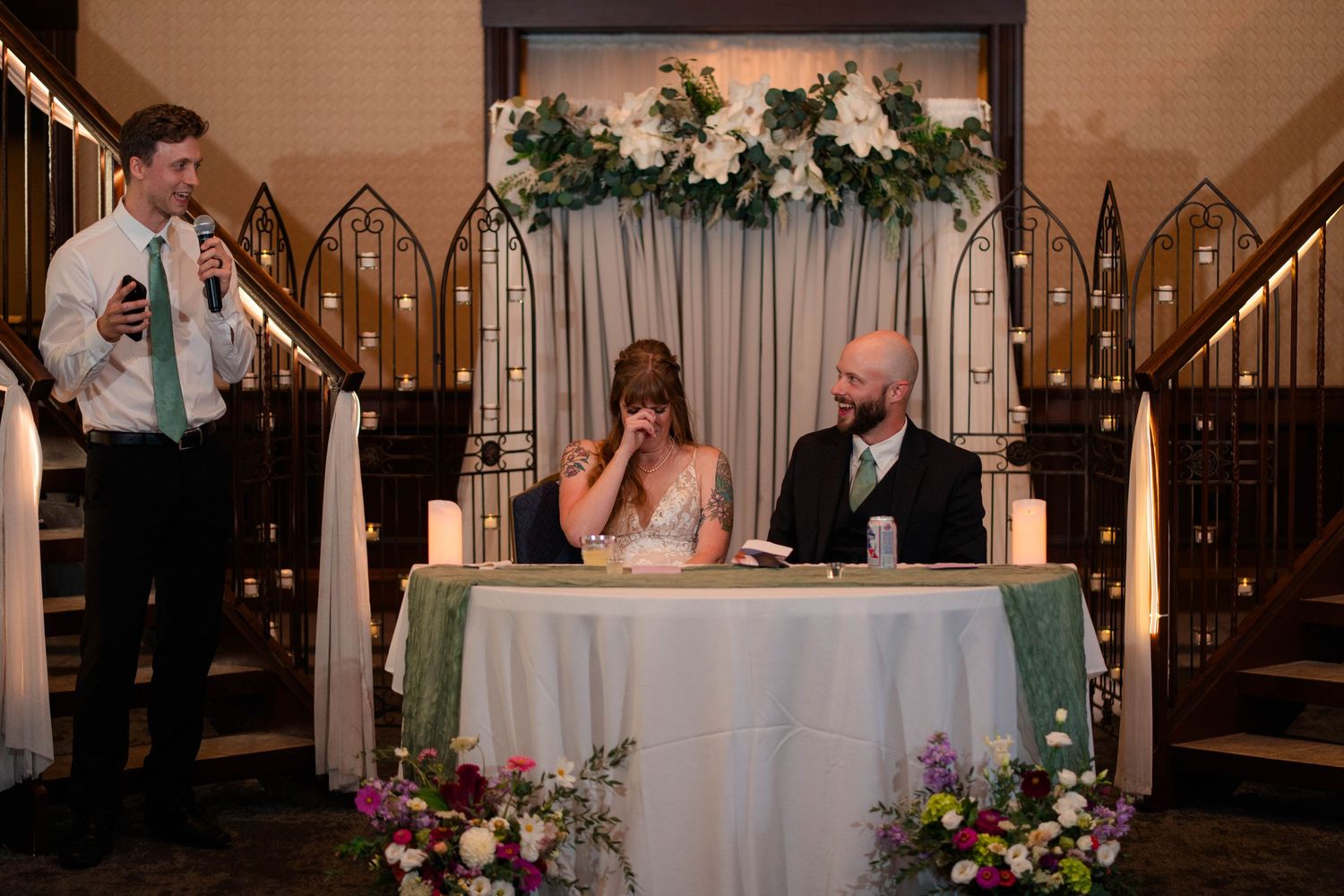 Bride laughs emotionally during wedding speech at elegant reception with floral arch backdrop.