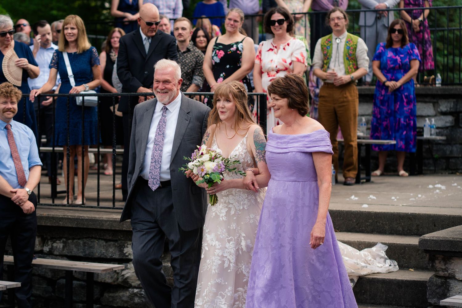 Bride in lace gown walks down aisle escorted by parents at outdoor wedding ceremony.