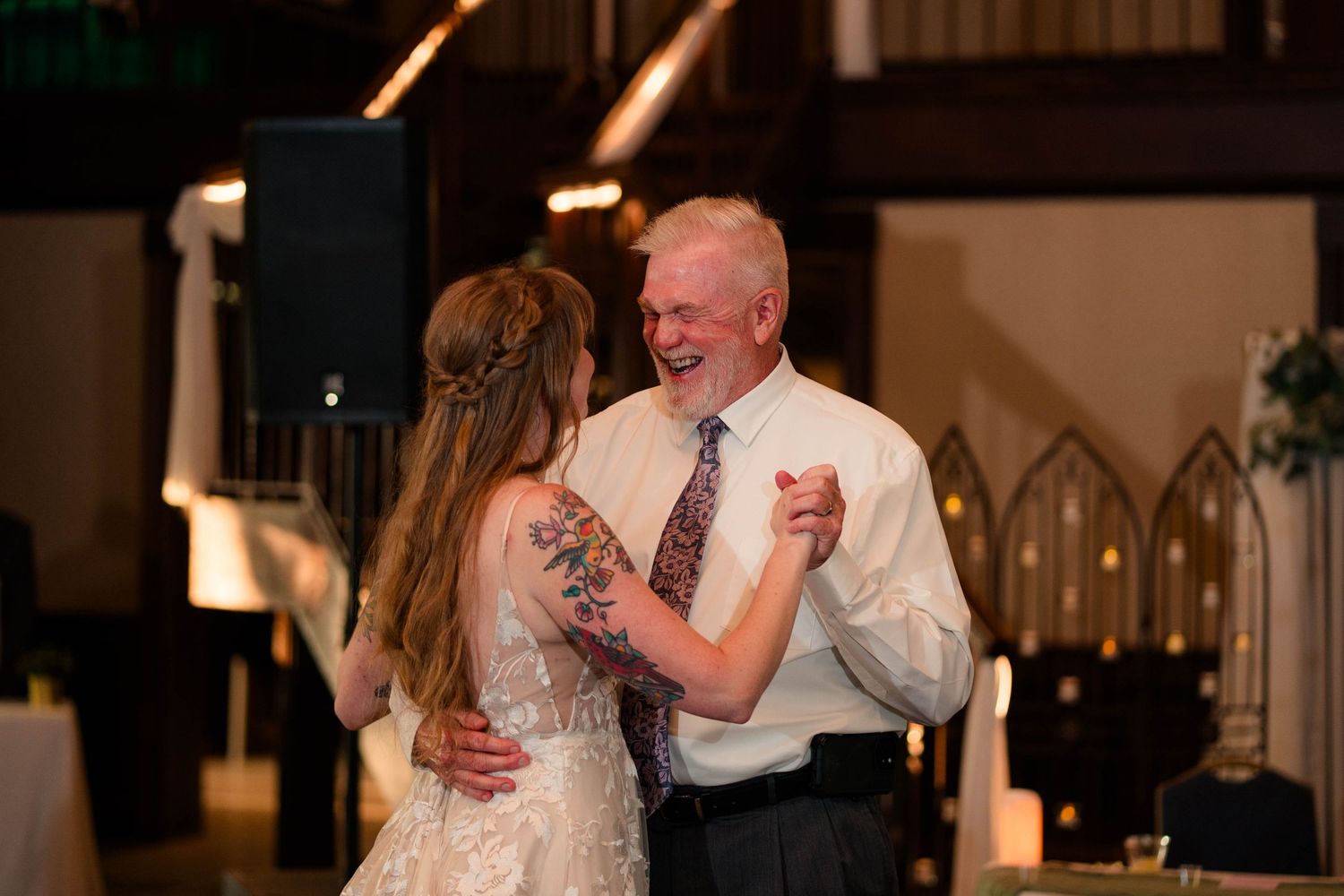 Bride in floral lace gown shares a joyful father-daughter dance inside a warmly lit reception venue.