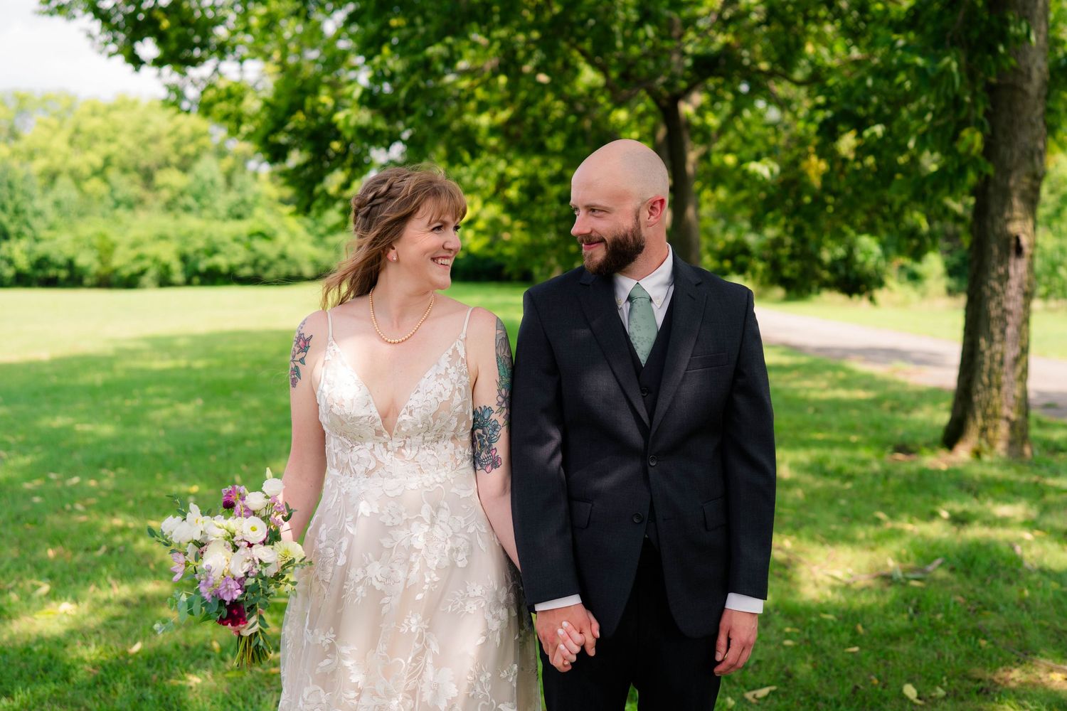 Bride and groom walk hand in hand through lush green park on their wedding day.