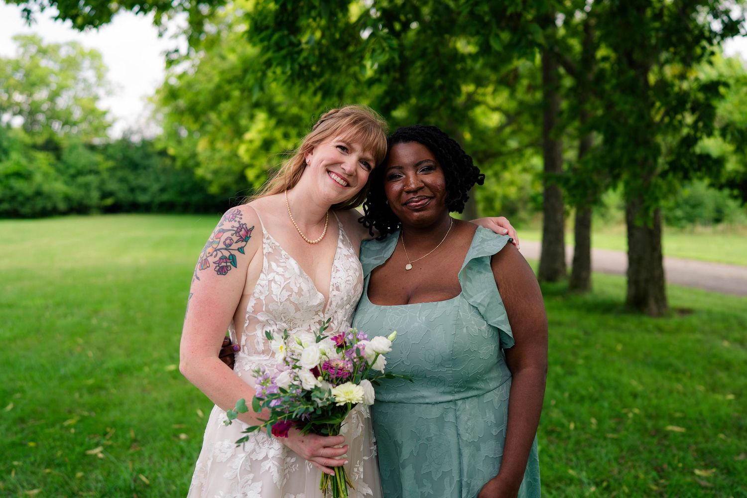 Bride in lace gown holding bouquet poses with bridesmaid in teal dress outdoors.