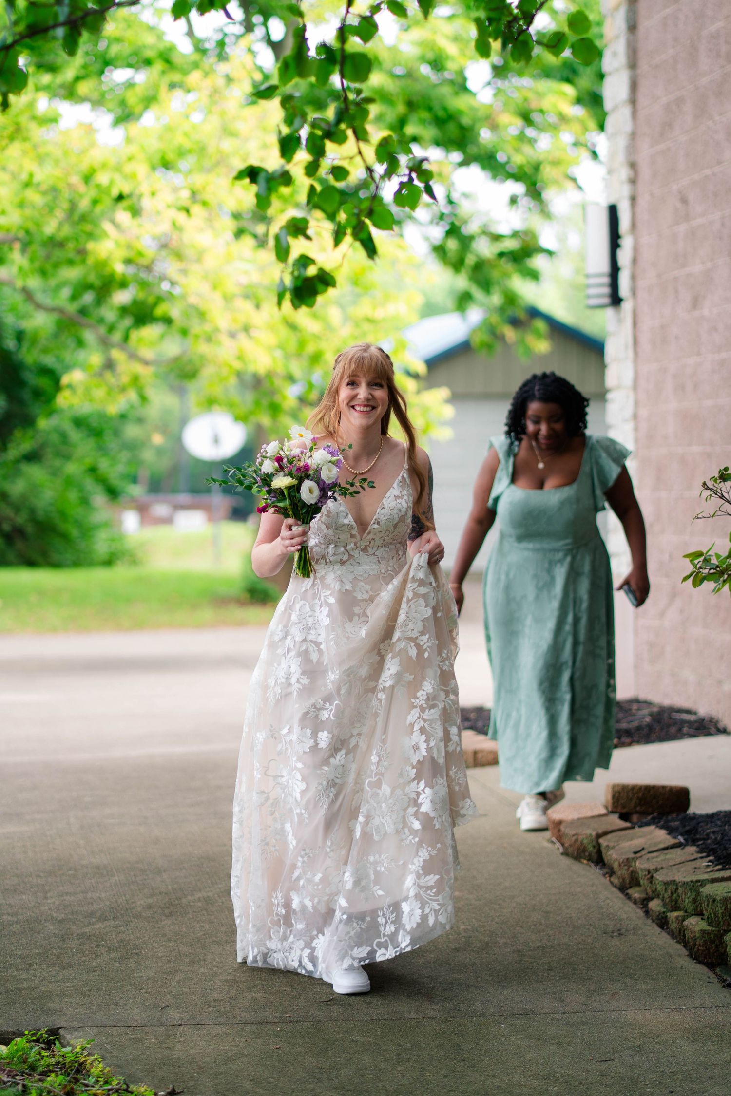 A smiling bride in a lace gown carries a bouquet while walking outdoors with a bridesmaid in sage green.