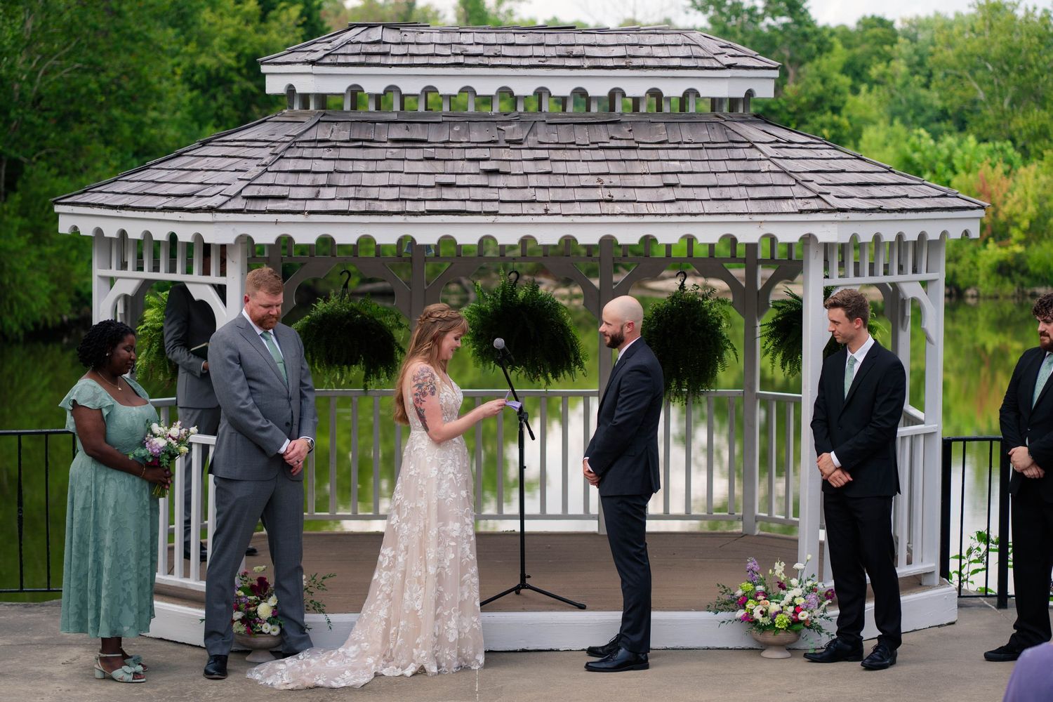 Wedding ceremony at white gazebo with bride and groom exchanging vows beside officiant and wedding party near pond.