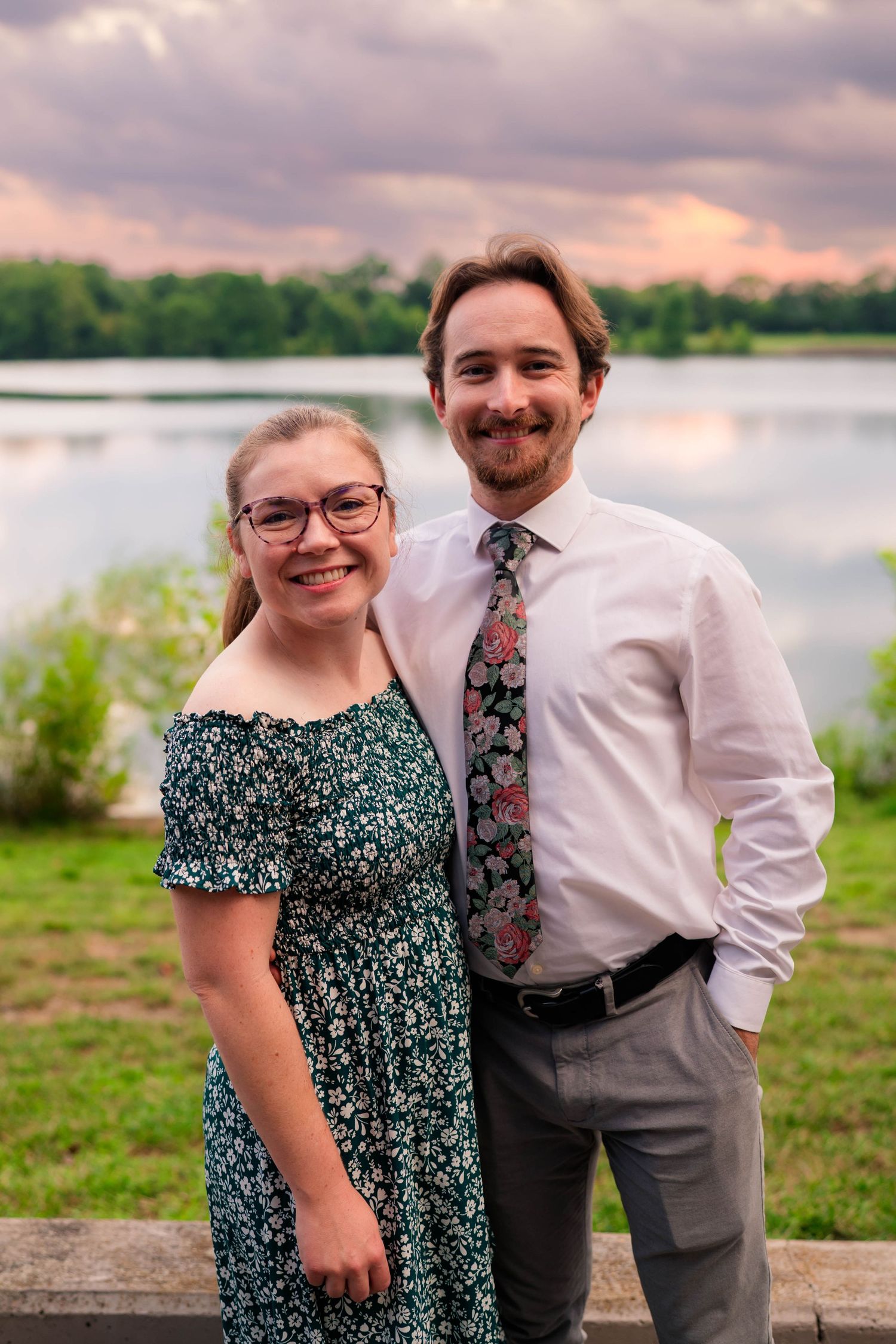 Smiling couple in floral tie and green dress poses together at sunset by a peaceful lake.