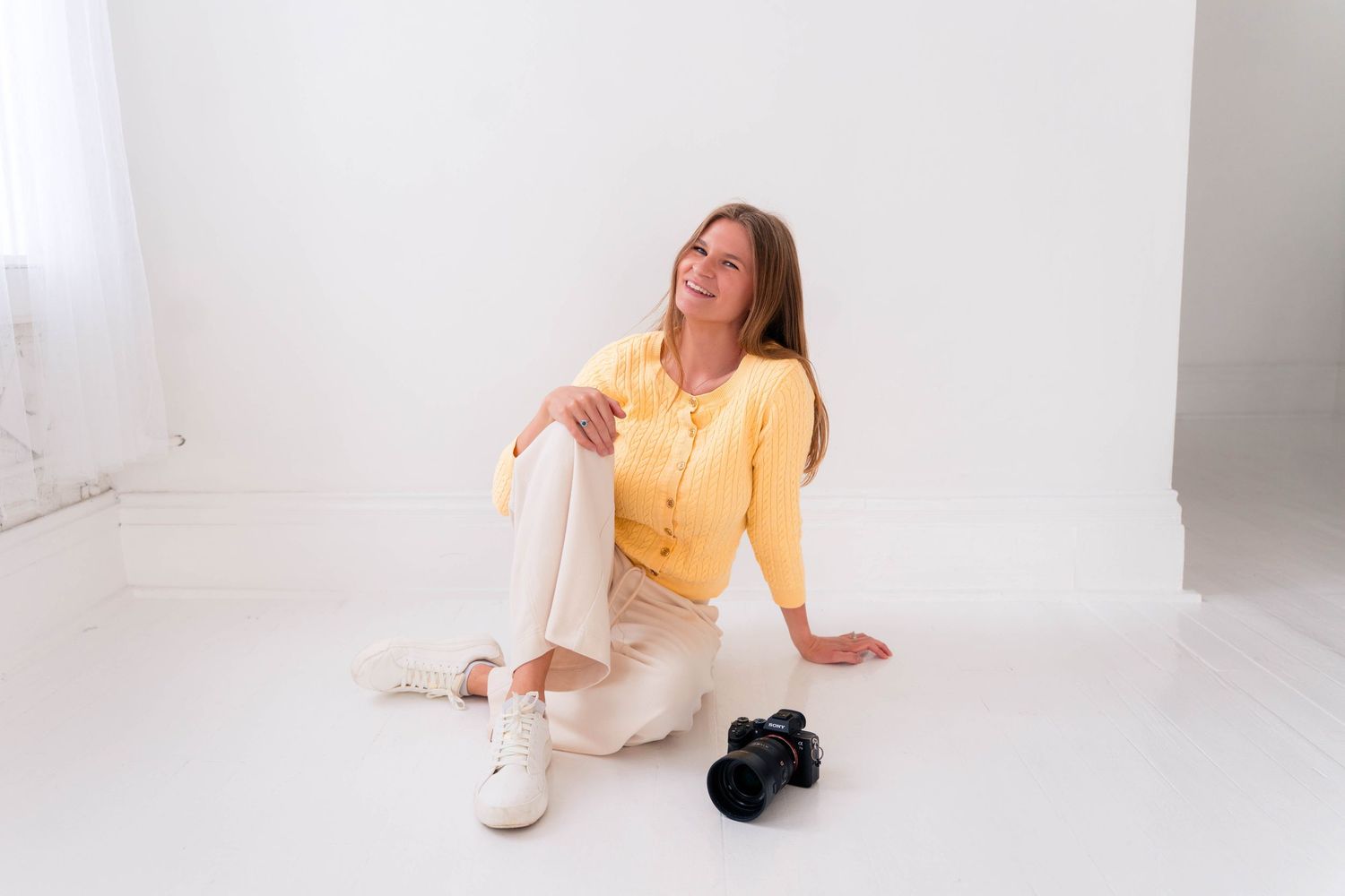 Female photographer in yellow cardigan sitting on white floor smiling with DSLR camera beside her.