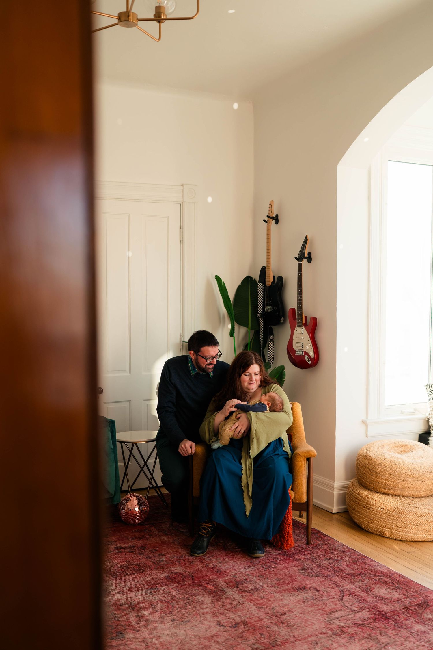 Couple sits together in cozy home with guitars on wall, tenderly holding their newborn baby.