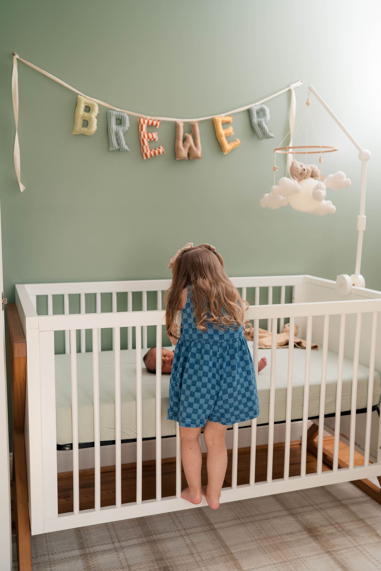 Toddler girl in blue dress standing at white crib beneath colorful BREWER name banner in nursery.