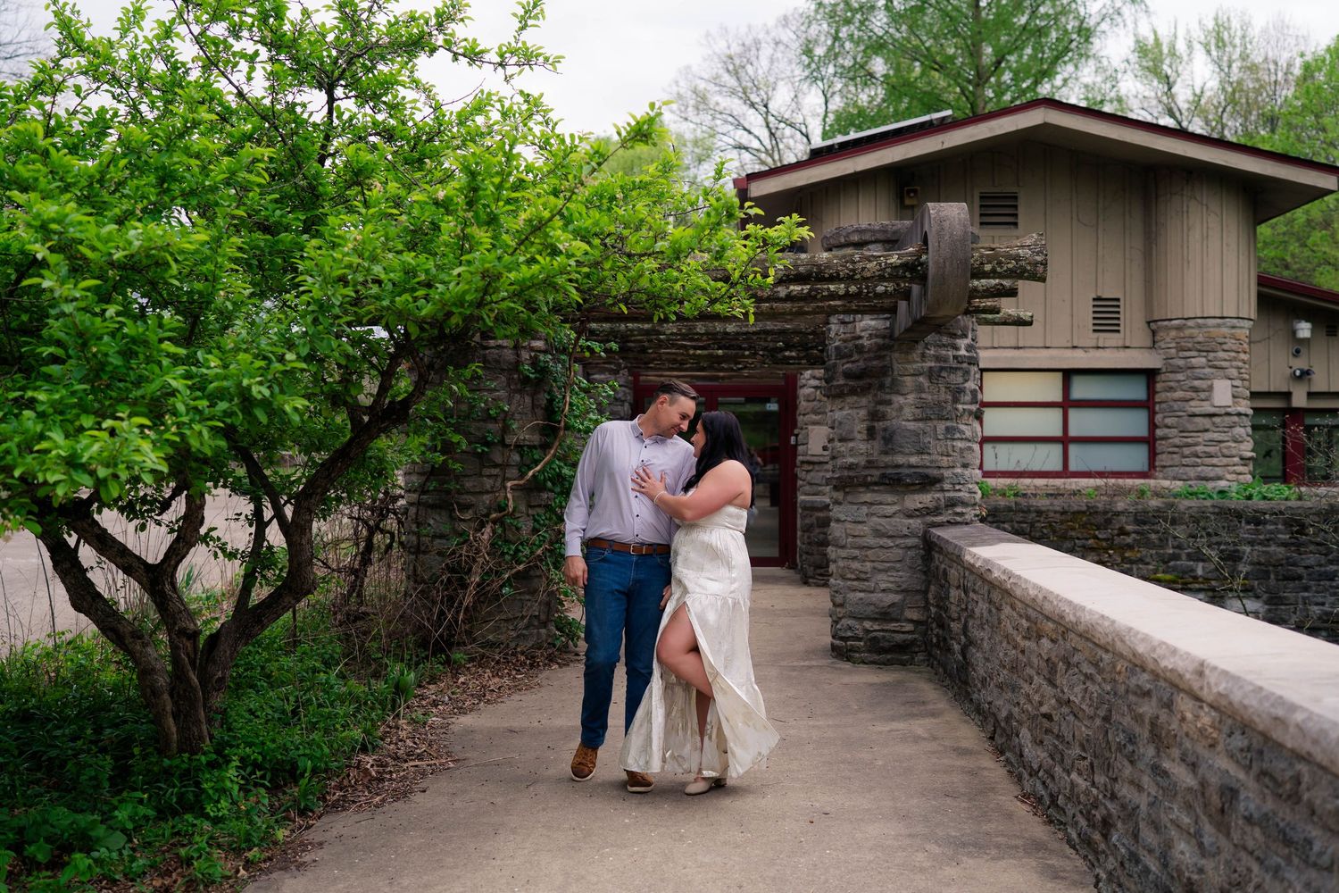 An engaged couple embraces on a stone bridge walkway near a craftsman-style building with spring greenery.