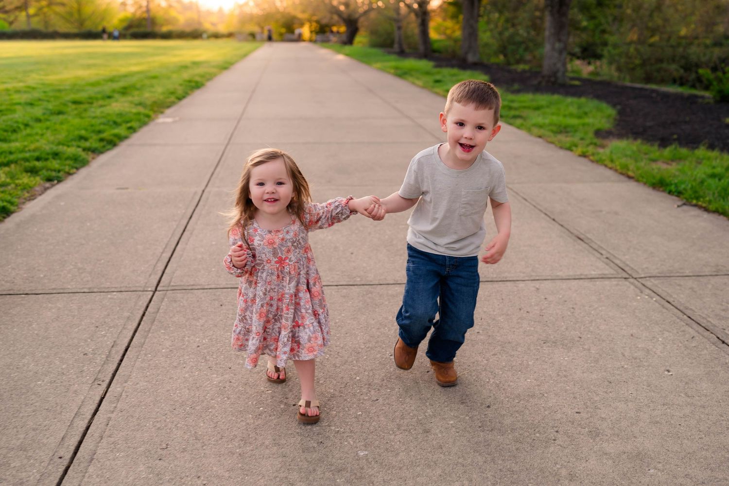 A young boy and girl in a floral dress hold hands while running joyfully down a sunny park pathway.