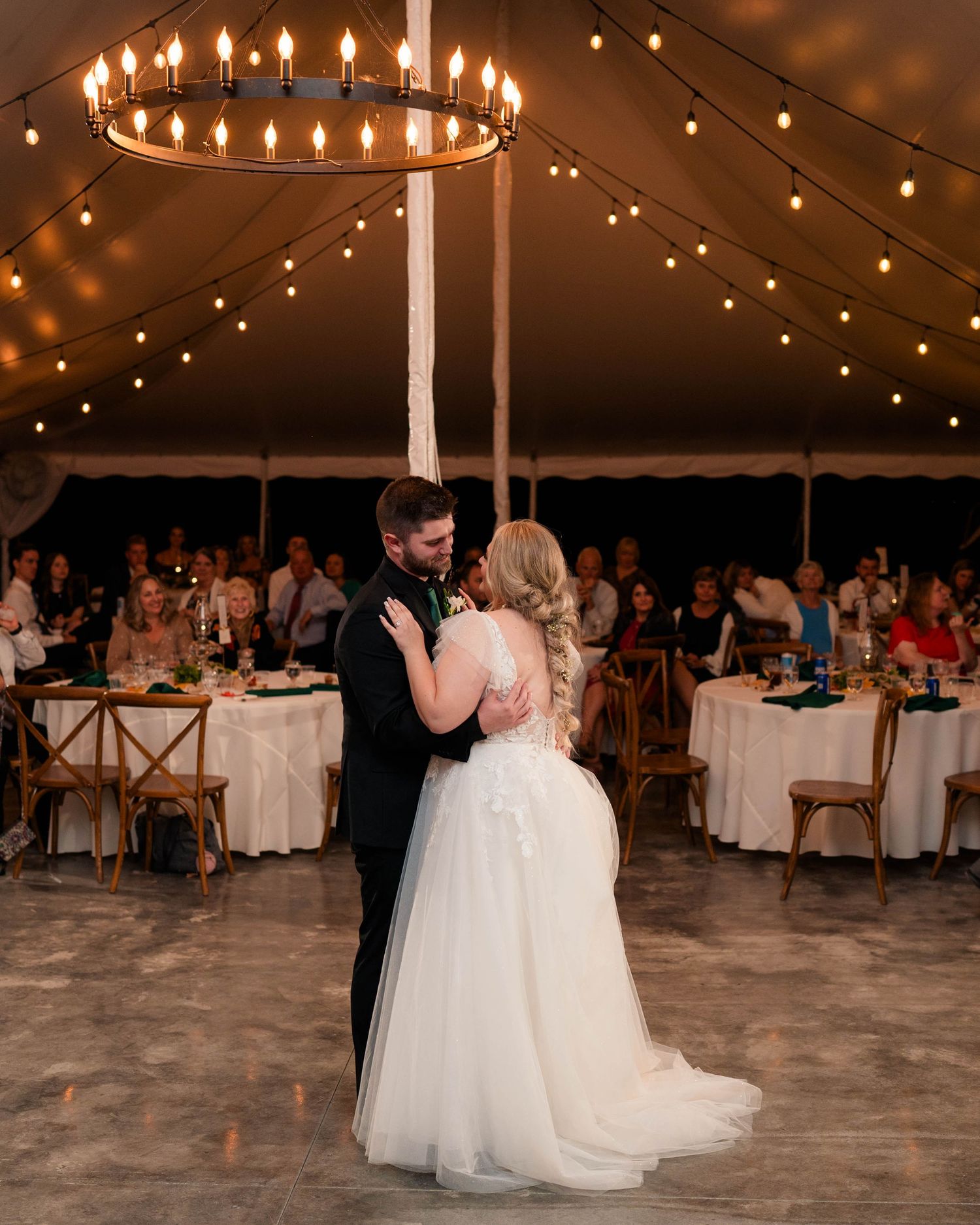 Bride and groom share first dance under chandelier and string lights in elegant tent wedding reception.