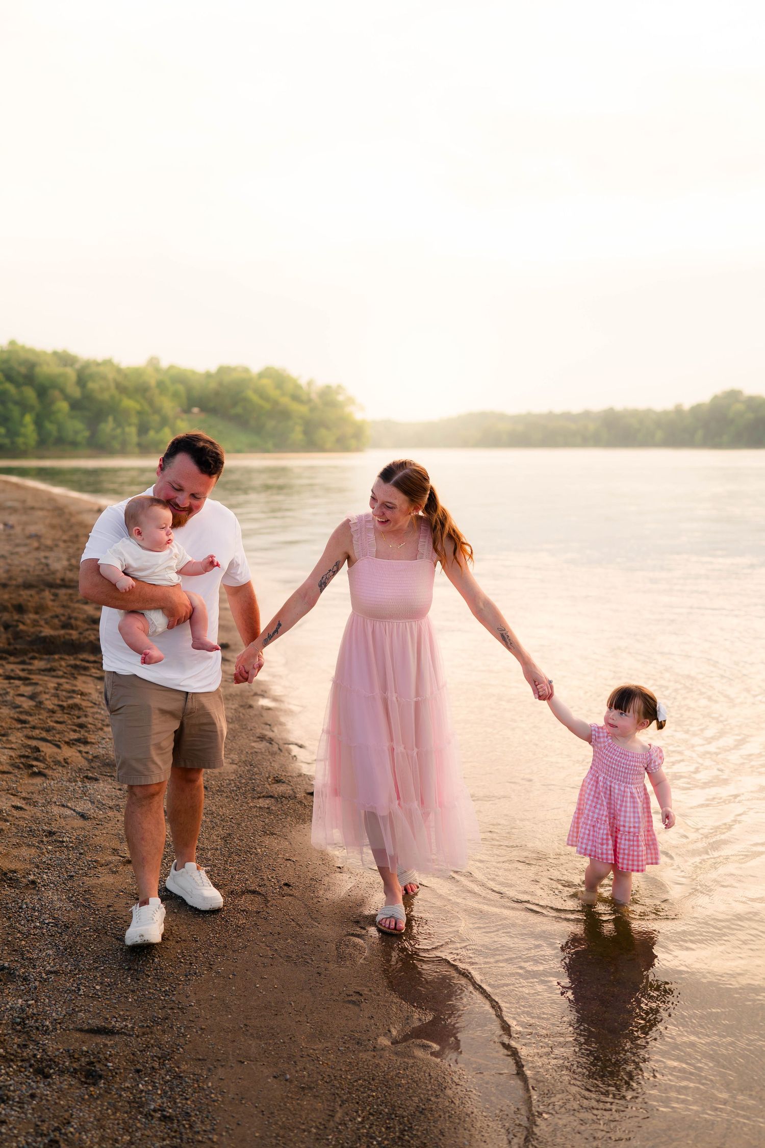 Happy family walking along a sandy beach at sunset, father holding baby, mother in pink dress holding toddler's hand.
