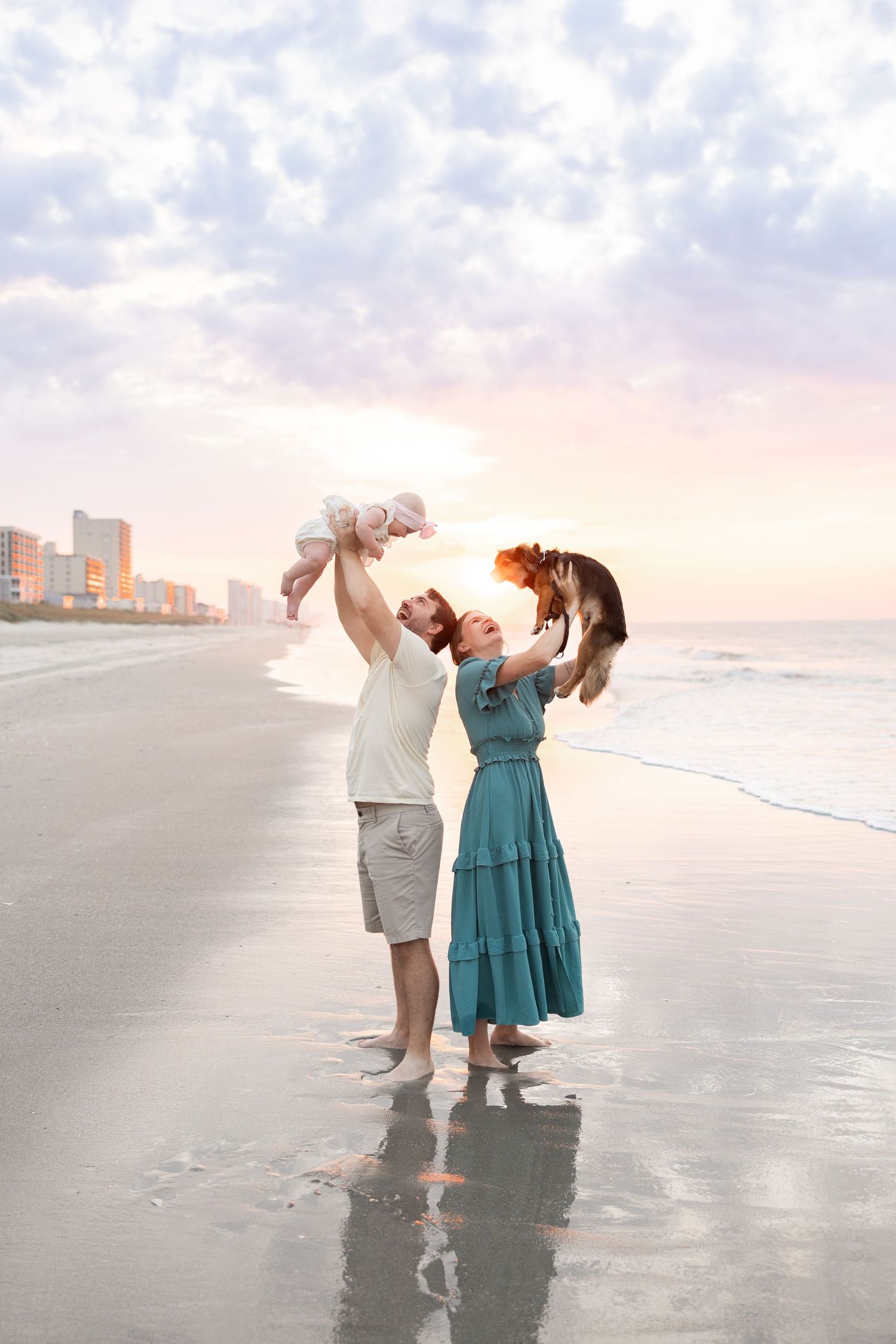 Couple lifting their baby and dog on a beach at sunset with ocean waves behind them.