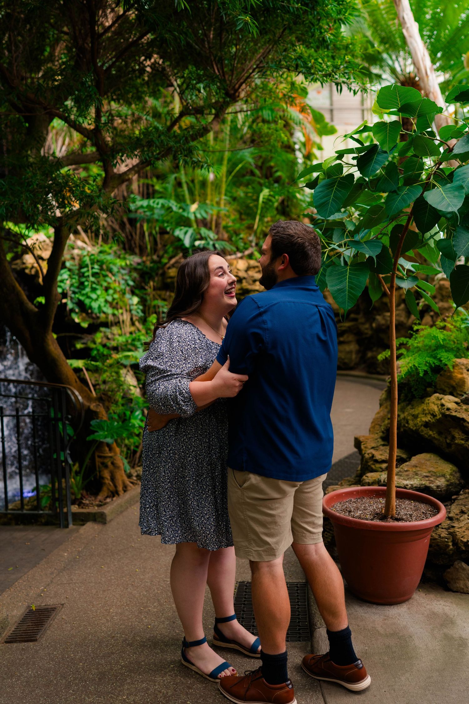A laughing couple embraces on a garden path surrounded by lush tropical greenery and a small waterfall.