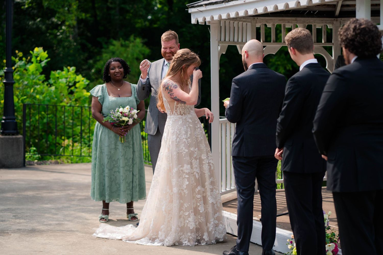 Bride and groom exchange vows at outdoor wedding ceremony near white gazebo with bridesmaid in green dress nearby.
