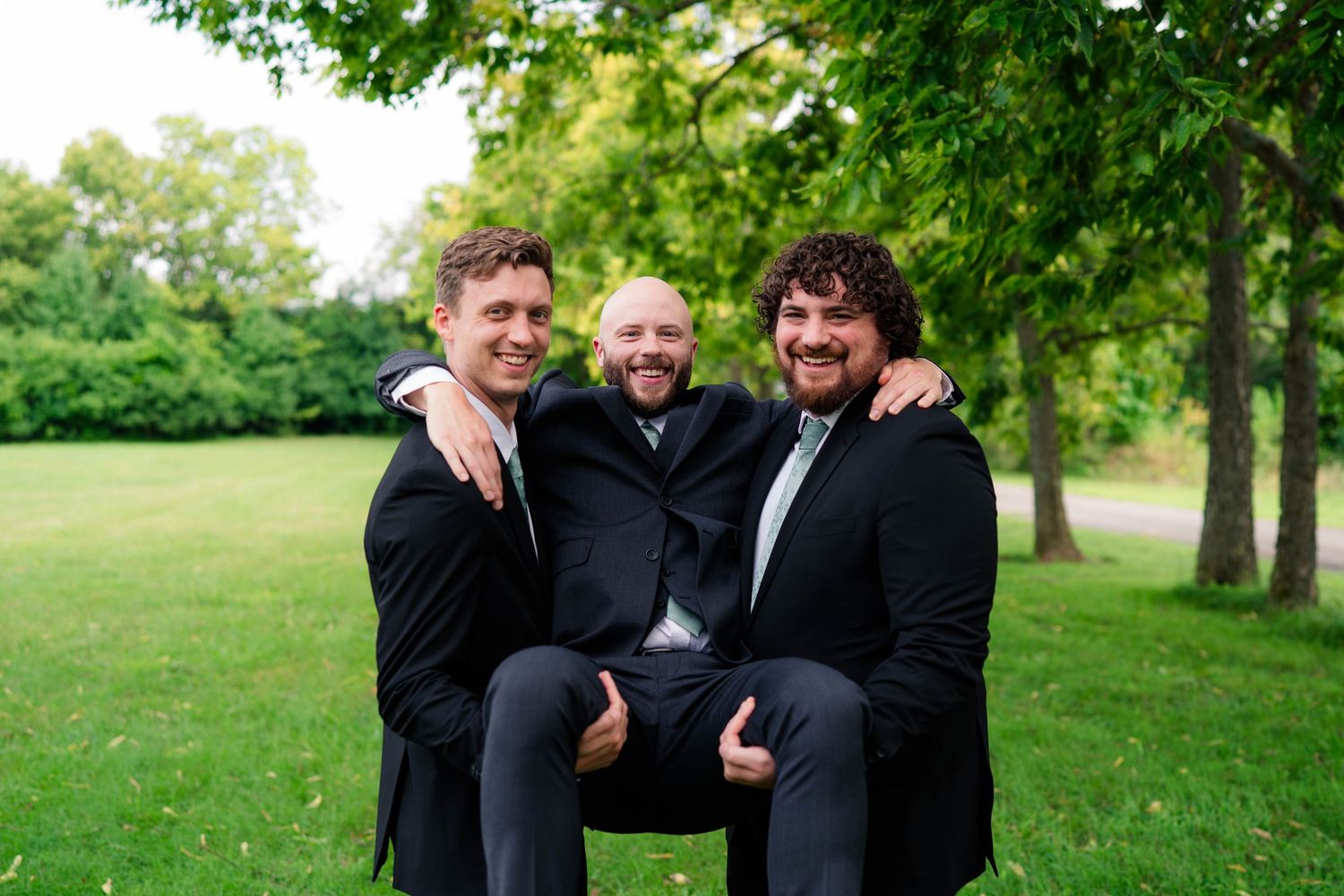 Two groomsmen playfully lift the groom in a park during wedding celebrations.