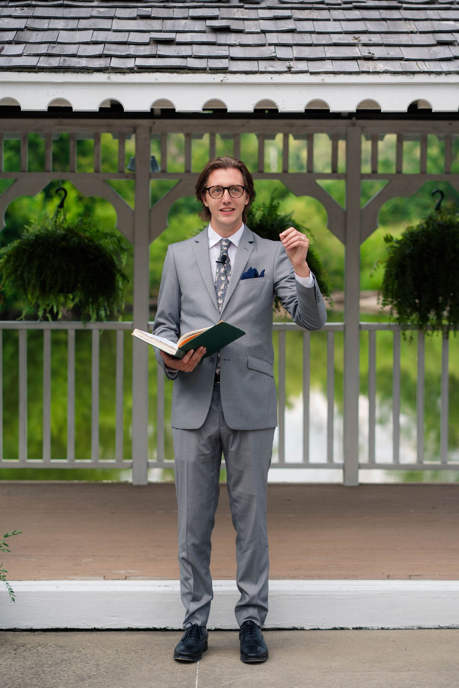 Wedding officiant in gray suit holding a book and pen while standing in front of a white gazebo.