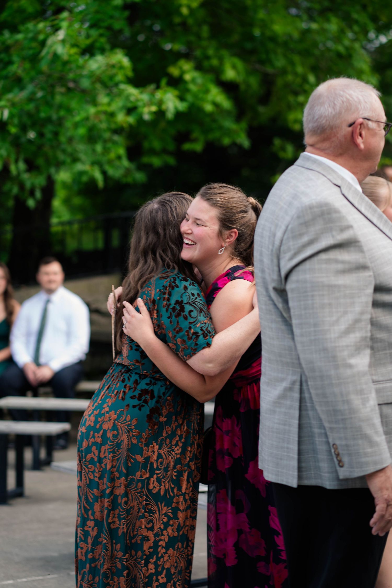 Two women share a warm embrace and smile at an outdoor wedding ceremony.