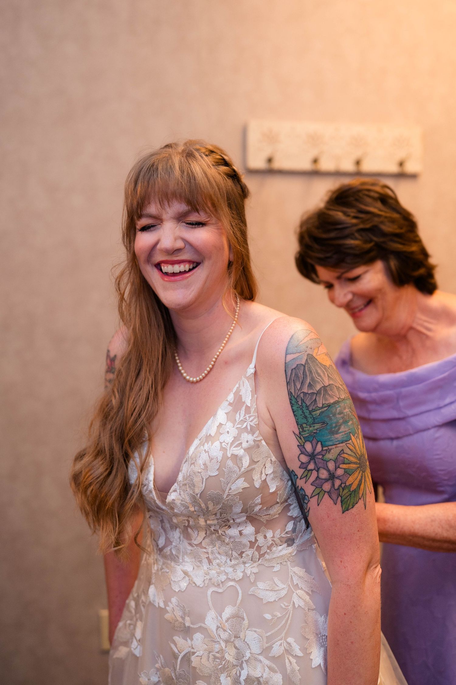 Laughing bride in lace wedding dress with floral tattoo and pearl necklace being helped by woman in lavender dress.