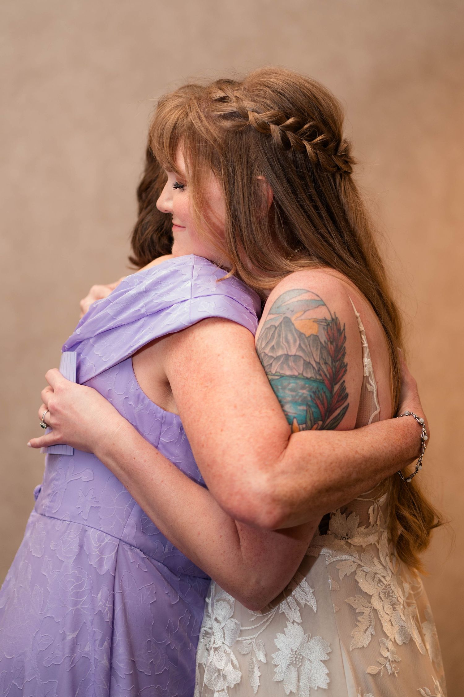 Bride in lace wedding dress with floral tattoo embraces woman in lavender dress at wedding.