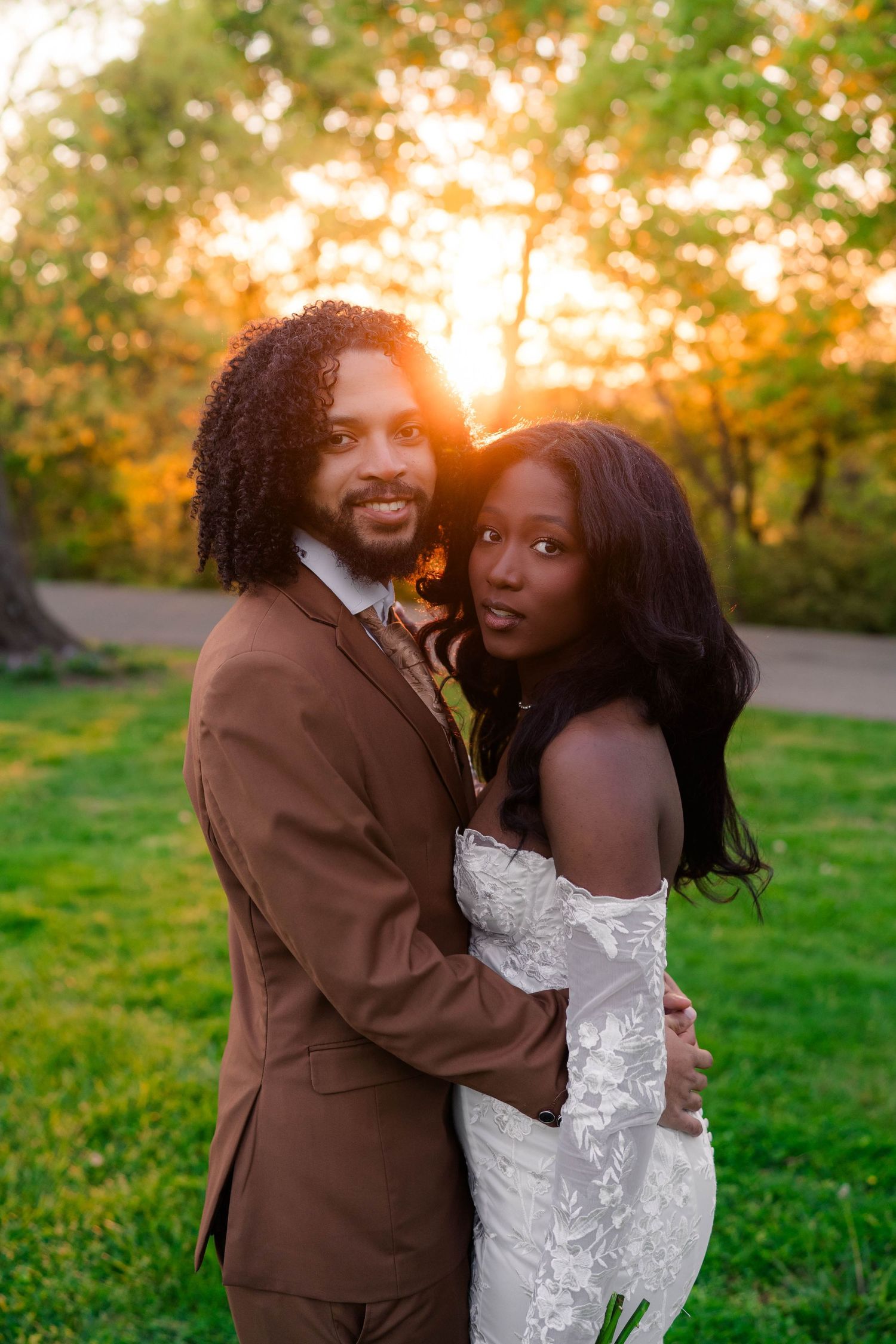 Newlyweds embrace at golden sunset in park, groom in brown suit, bride in off-shoulder lace wedding gown.