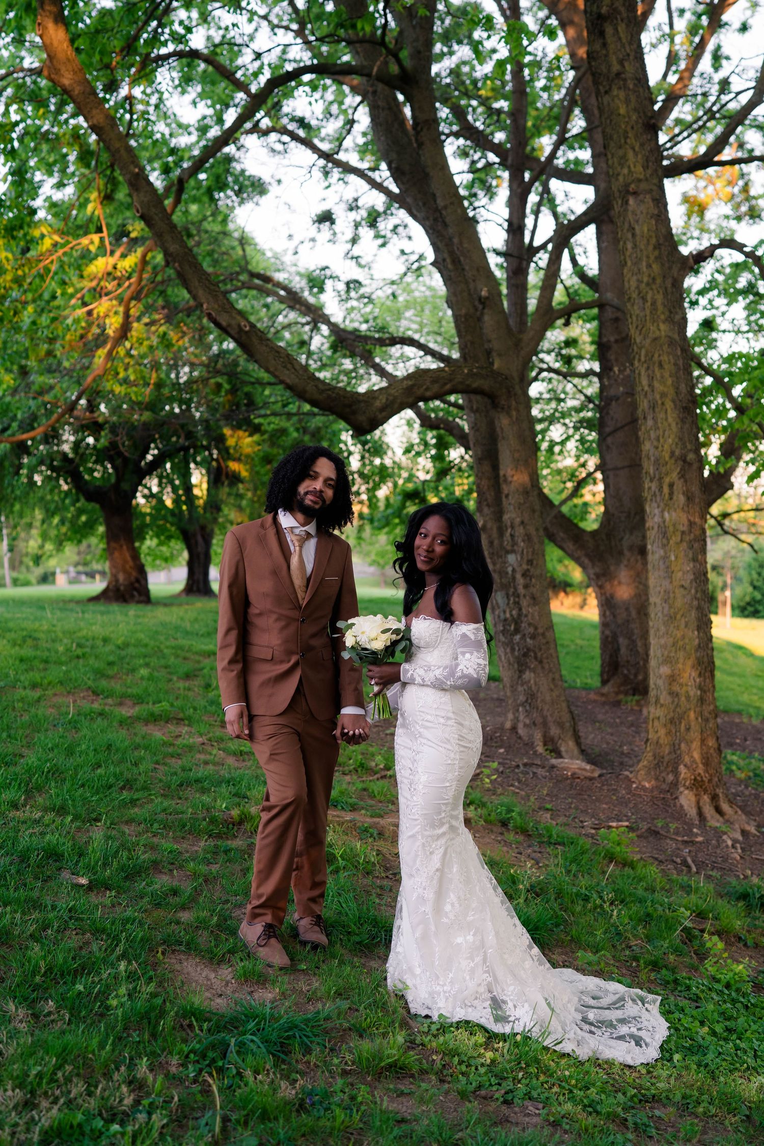 Bride and groom hold hands walking through lush green park, bride in lace gown, groom in brown suit.