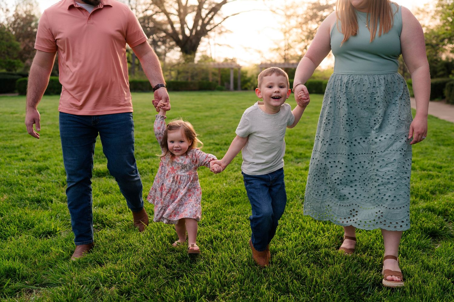 Family of four walking hand-in-hand on green grass at sunset, two young children laughing joyfully.