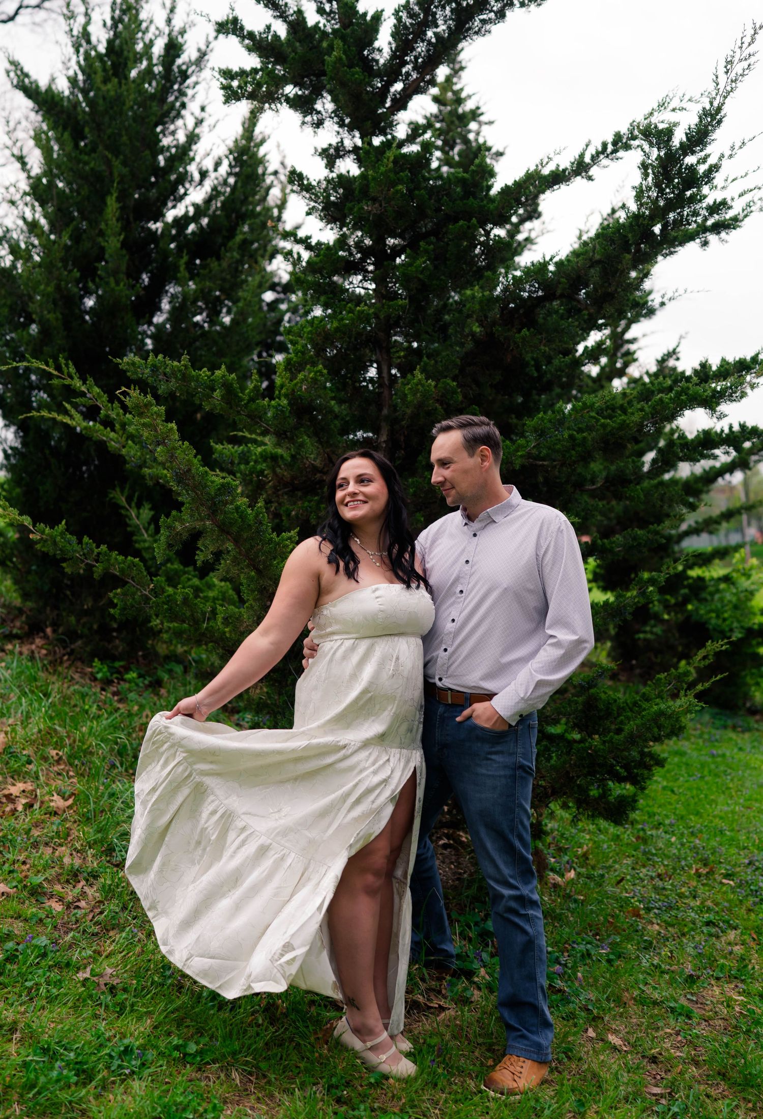 Smiling couple posing together near evergreen tree, woman in flowing white dress and man in gray shirt and jeans.