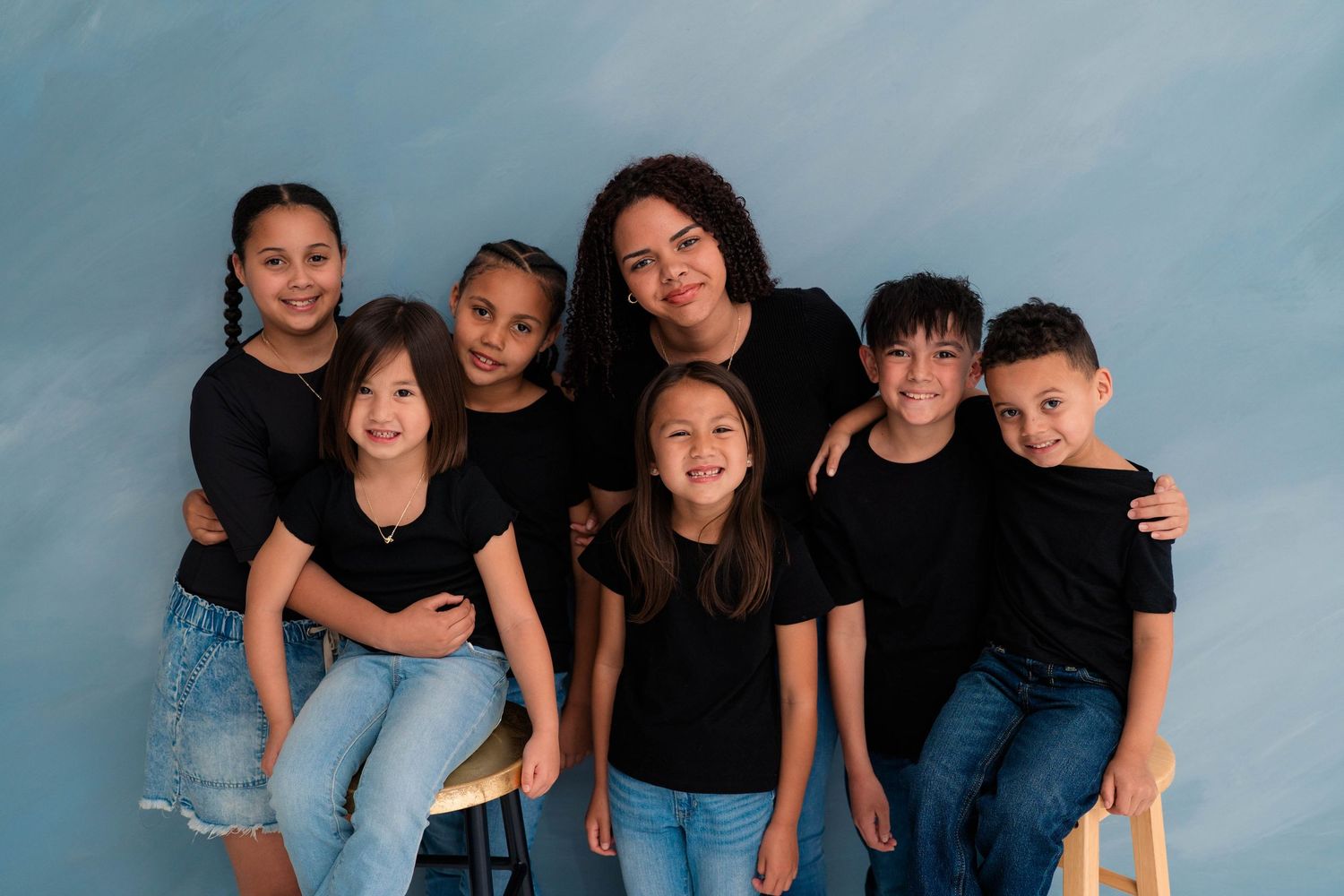 A woman poses with six children in matching black shirts against a blue background in a professional family portrait.