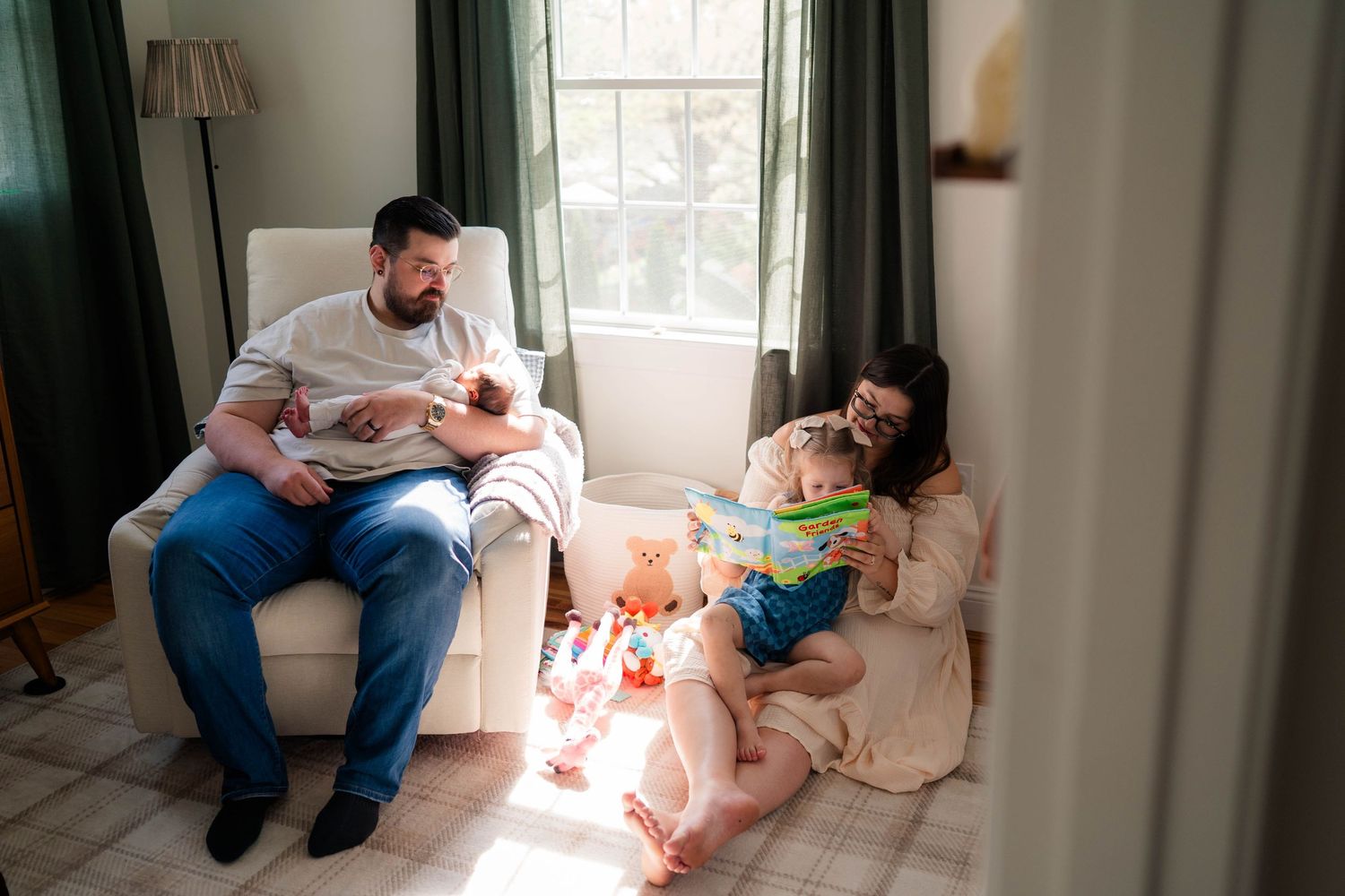 A family relaxes in a sunlit nursery, with dad holding a newborn and mom reading to their toddler.