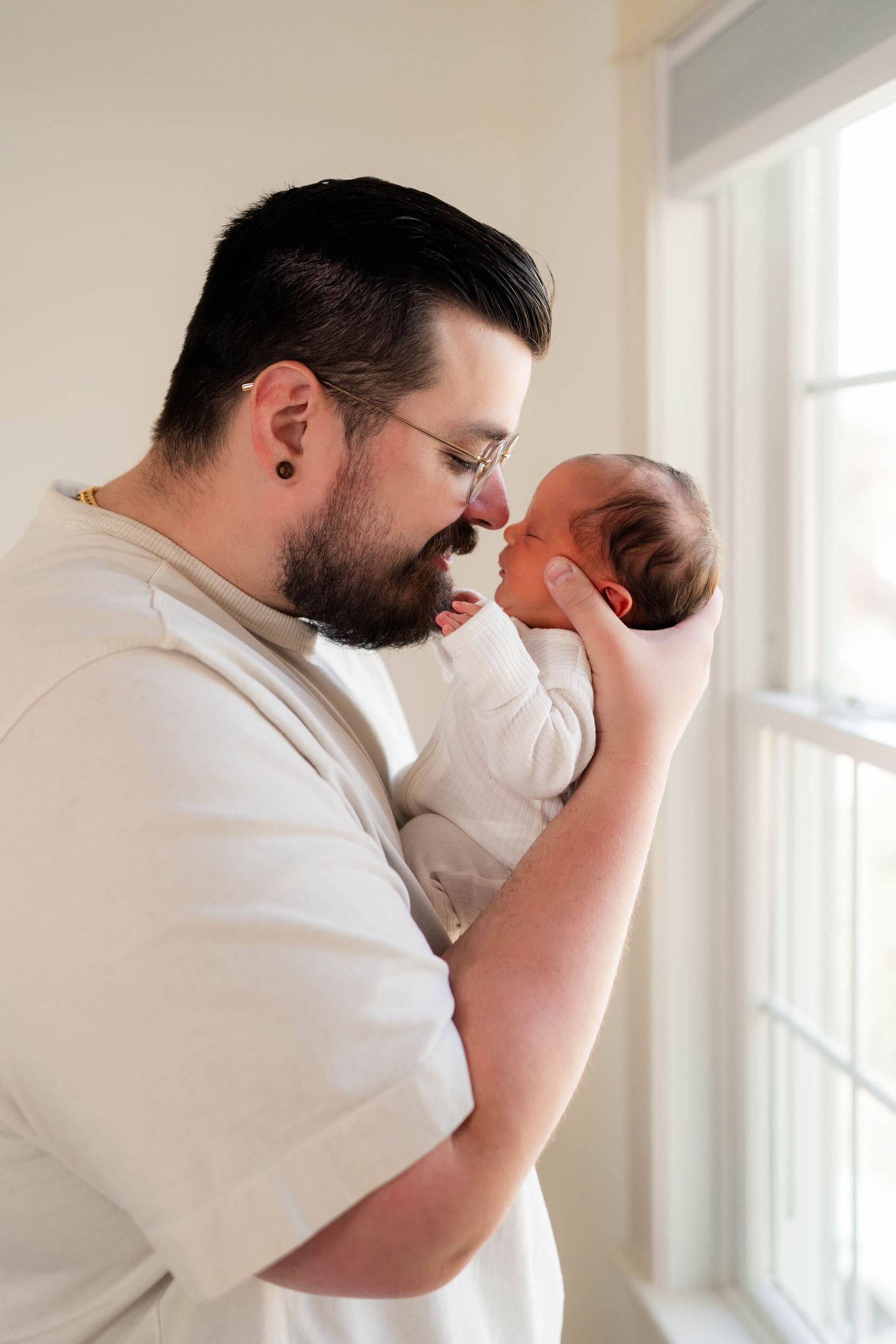 Loving father touches foreheads with his newborn baby while holding them gently near a window.