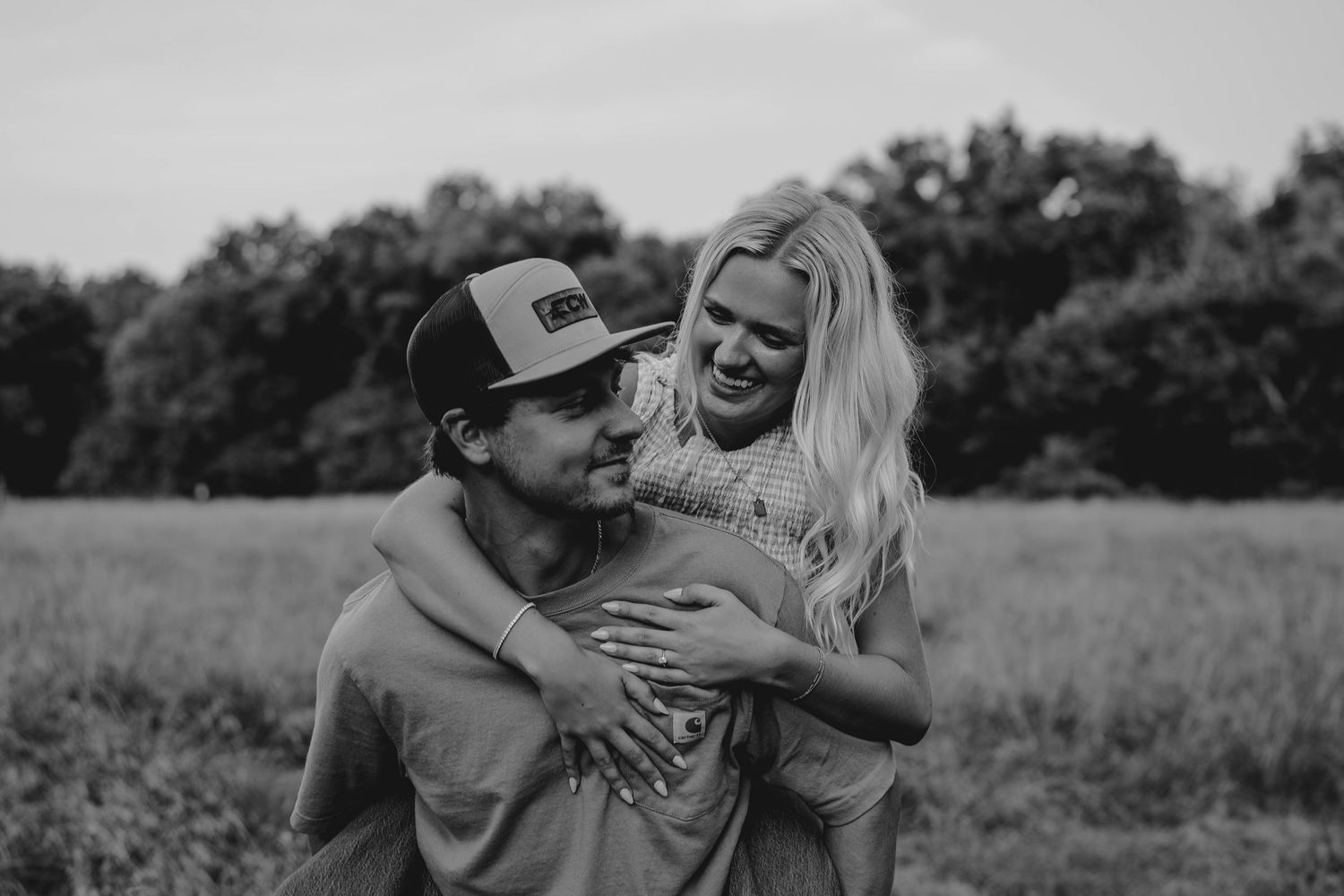 Black and white photo of a laughing couple embracing in a field, woman hugging man from behind.