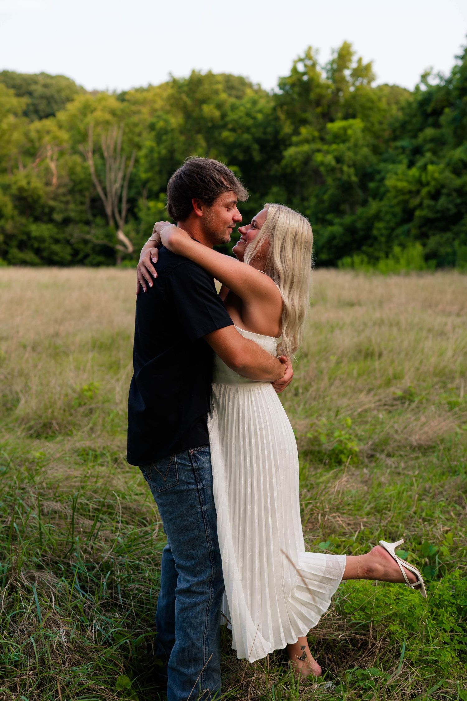 Couple embracing in a grassy field, woman in white dress and man in black shirt and jeans, with green trees behind.