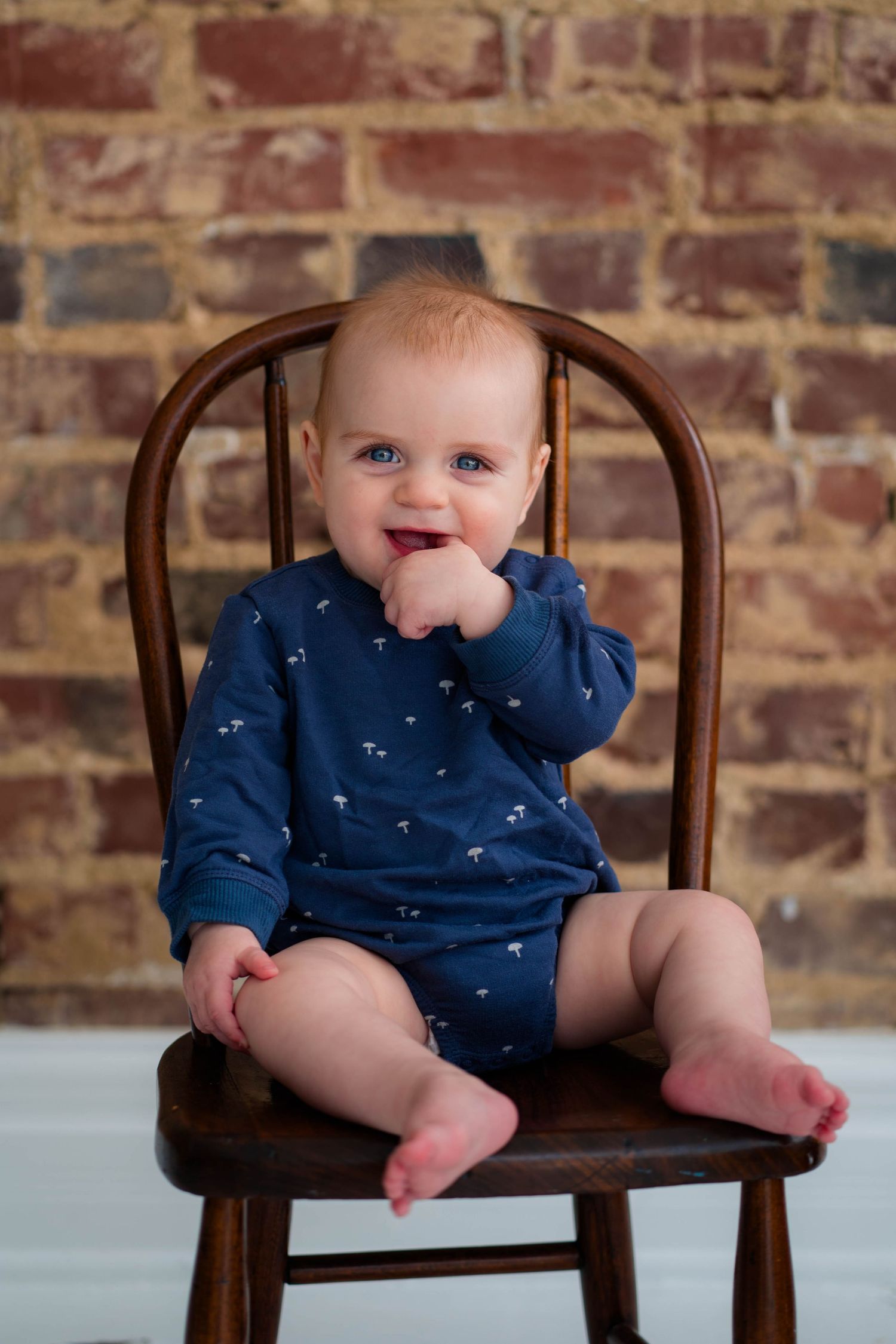 Smiling baby with blue eyes in navy romper sitting on wooden chair against rustic brick wall background.