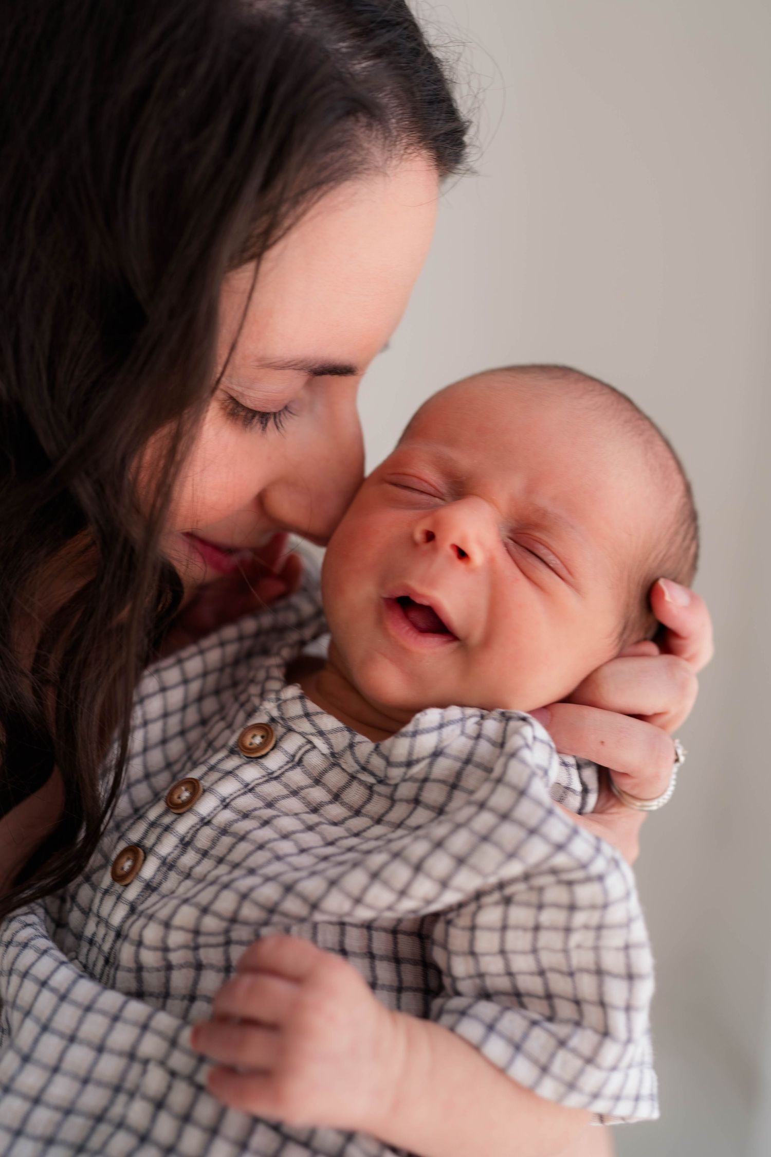 Mother tenderly kissing smiling newborn baby in plaid outfit against soft gray background.