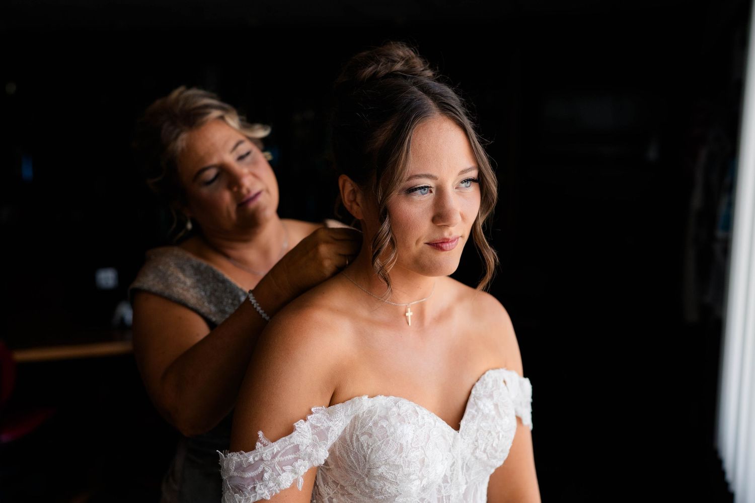 Mother helping bride with jewelry as she prepares in elegant off-shoulder lace wedding dress.