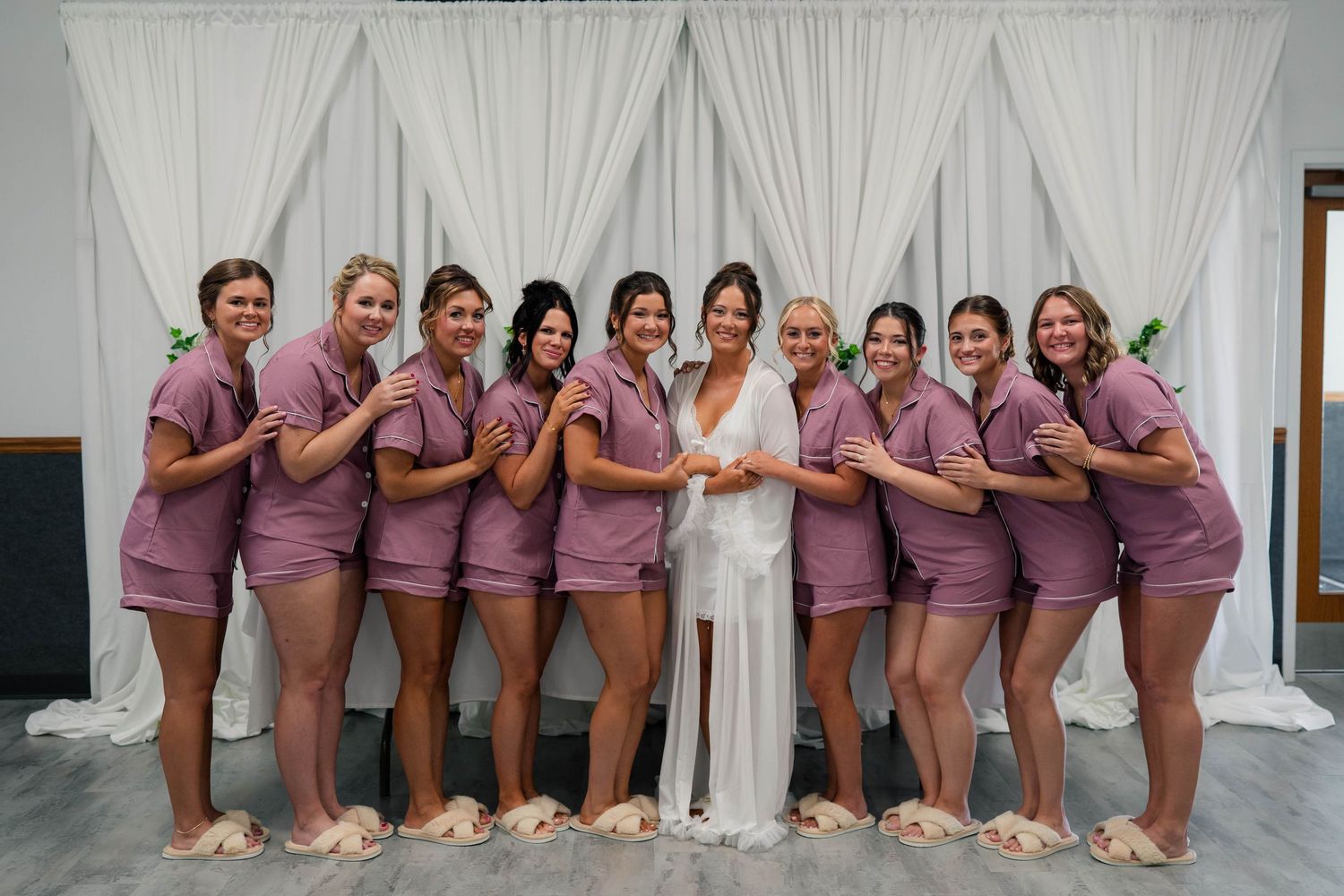 Bride in white robe poses with bridesmaids in matching mauve pajama sets forming a heart shape in front of white drape backdrop.