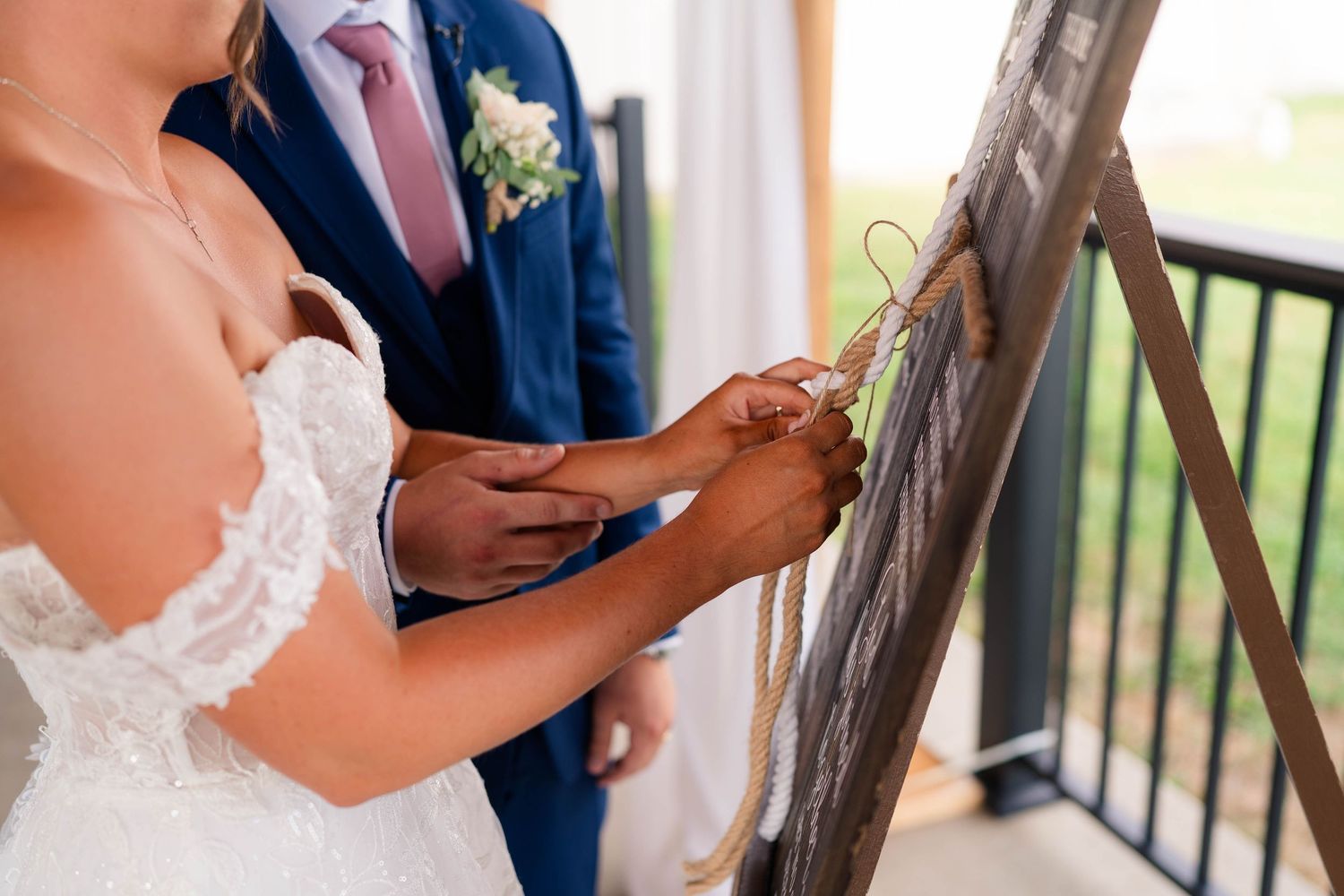 Bride and groom in navy suit participate in cord tying unity ceremony on outdoor balcony during wedding.
