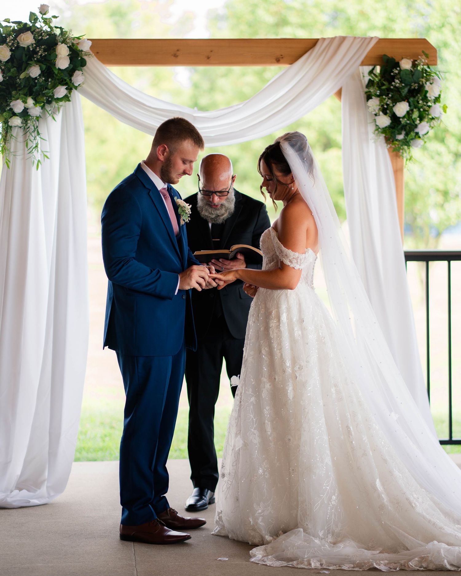 Groom places ring on brides finger under white draped arch at wedding ceremony.