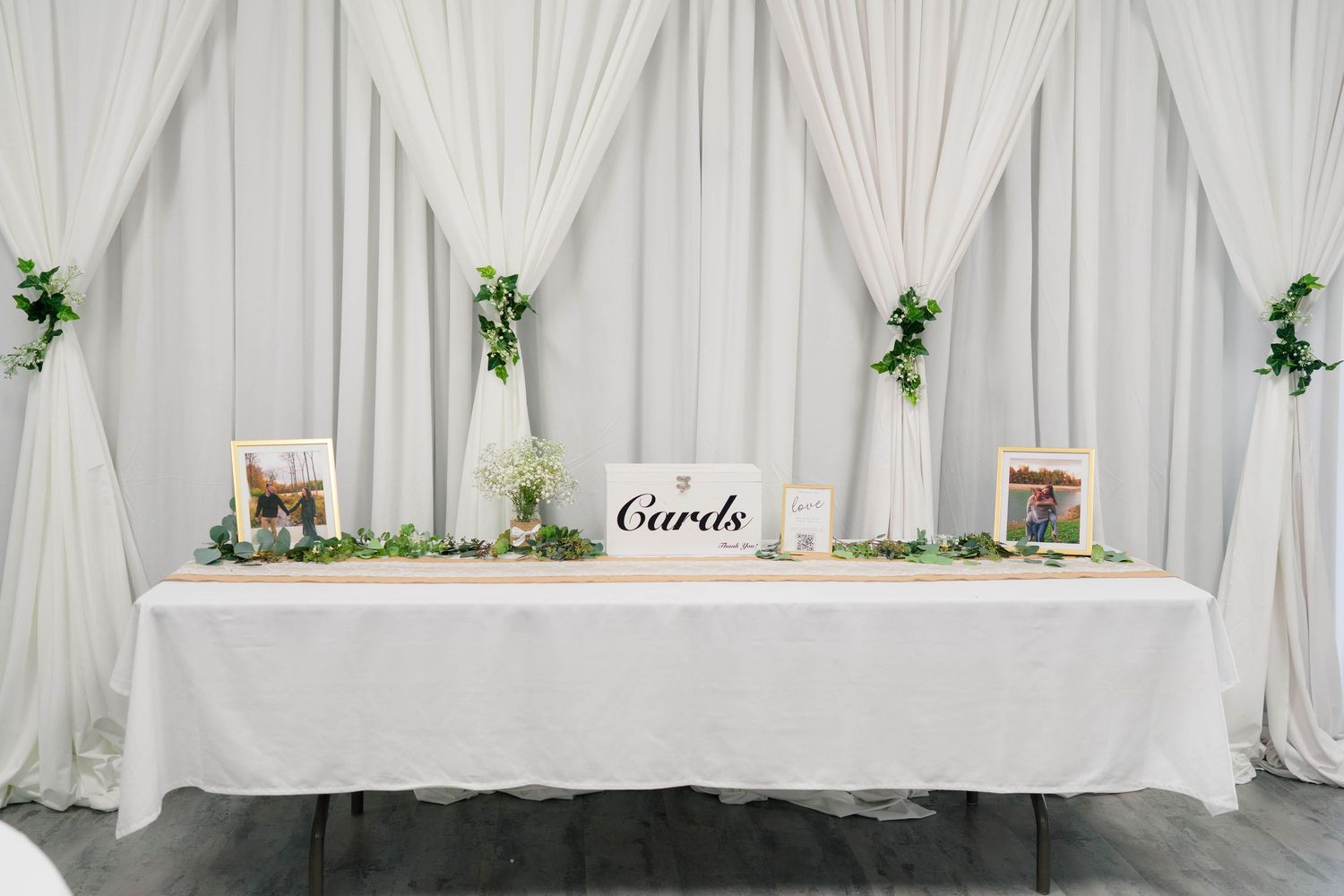 Elegant wedding cards table with white draping backdrop, greenery garland, framed photos, and Cards sign.