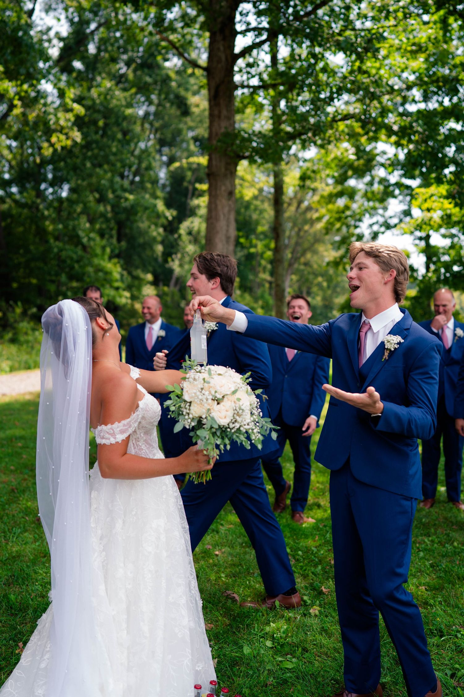 Joyful wedding party celebrating with bride and groomsmen in navy suits outdoors among trees.