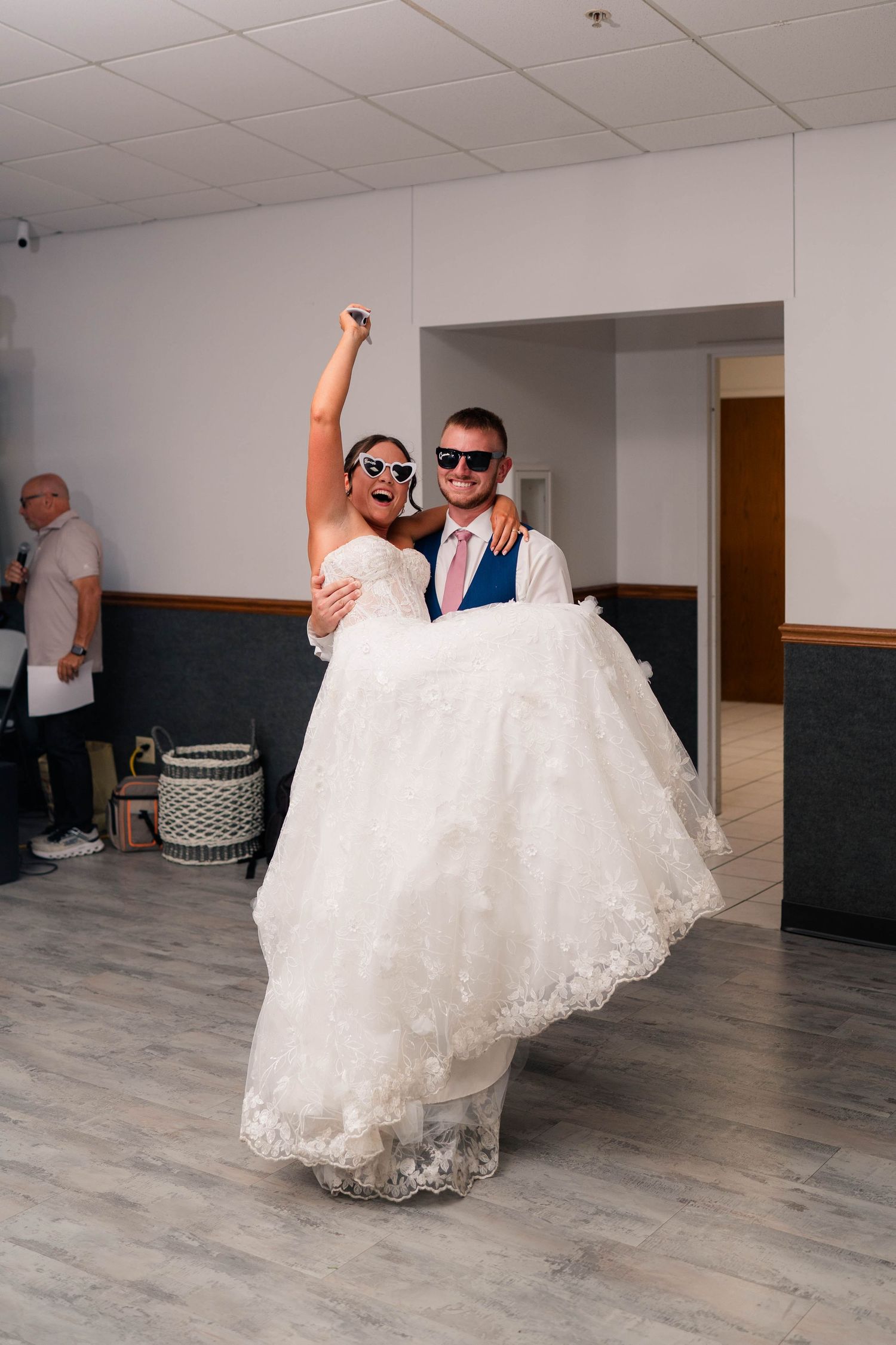 Groom in navy suit carries bride in white lace gown while both wear novelty sunglasses on the dance floor.