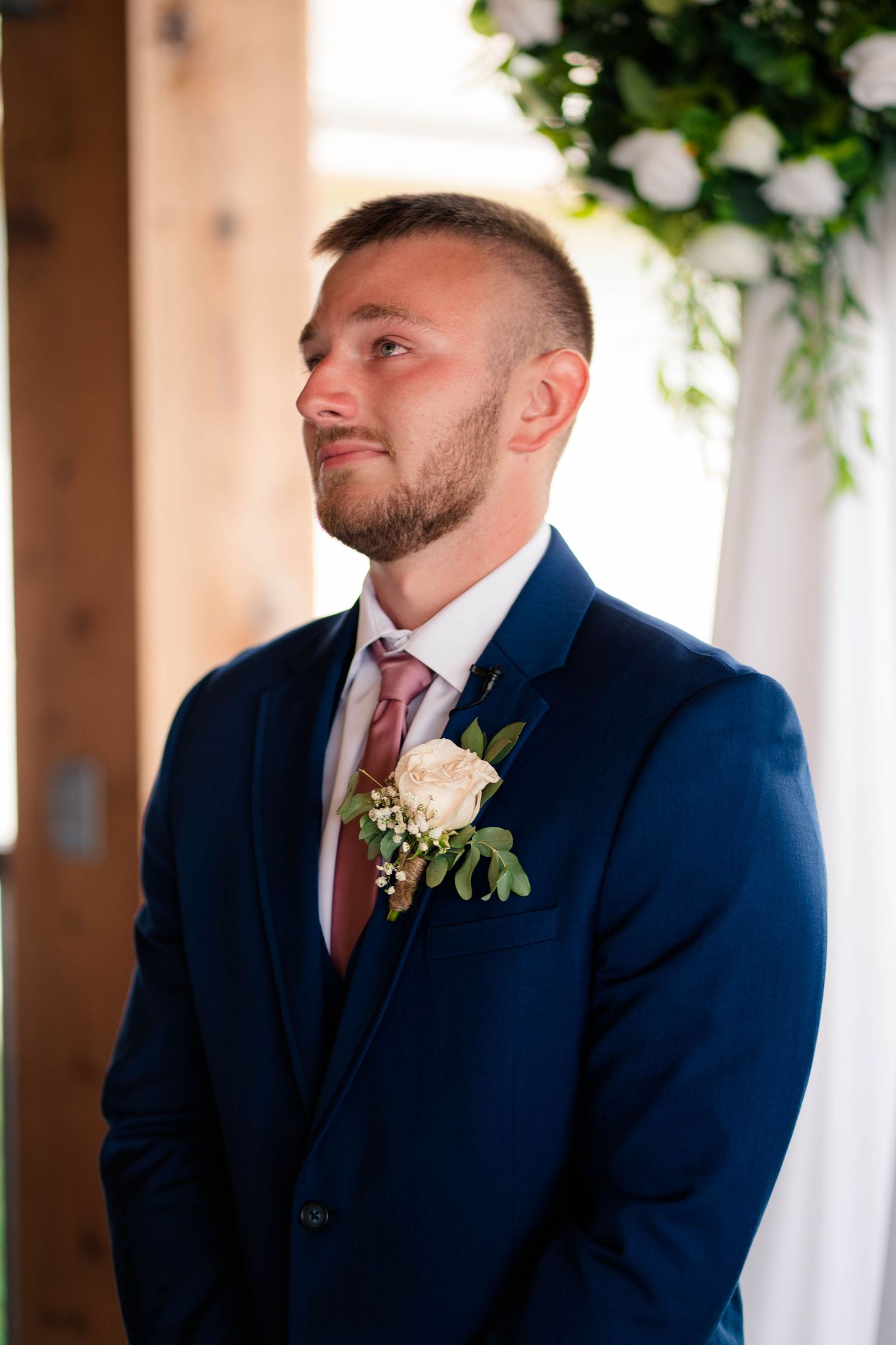 Emotional groom in navy suit with rose boutonniere watching bride walk down the aisle.