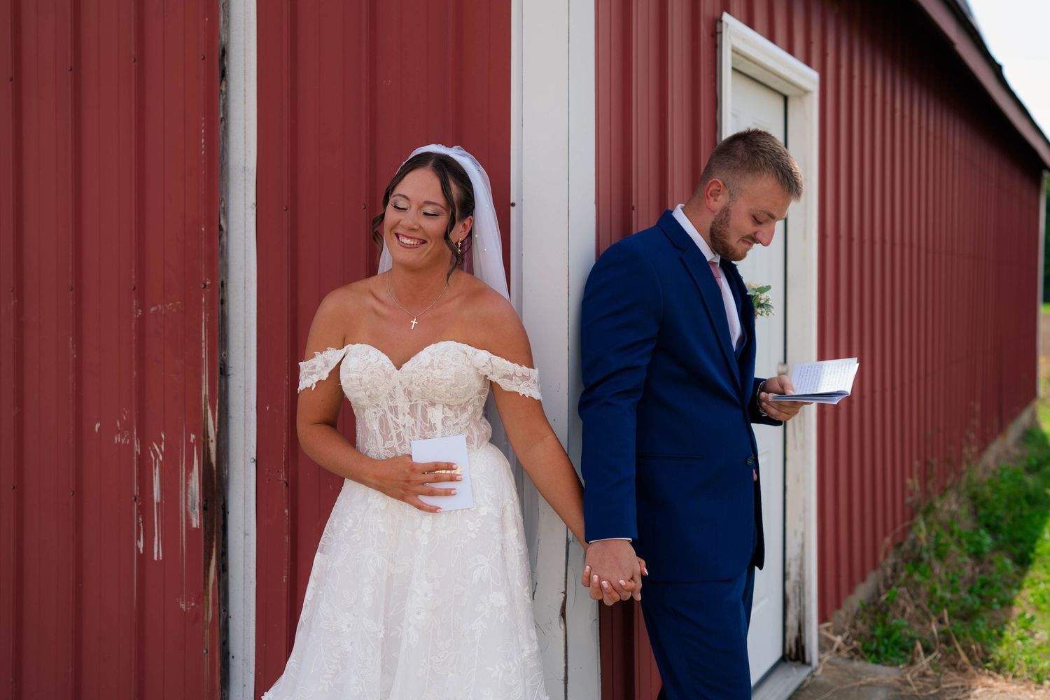 A smiling bride and groom hold hands while leaning against a red barn, groom reads a note.