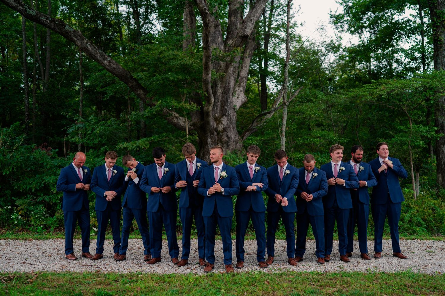 Groomsmen in navy blue suits with pink ties lined up outdoors before wedding ceremony.