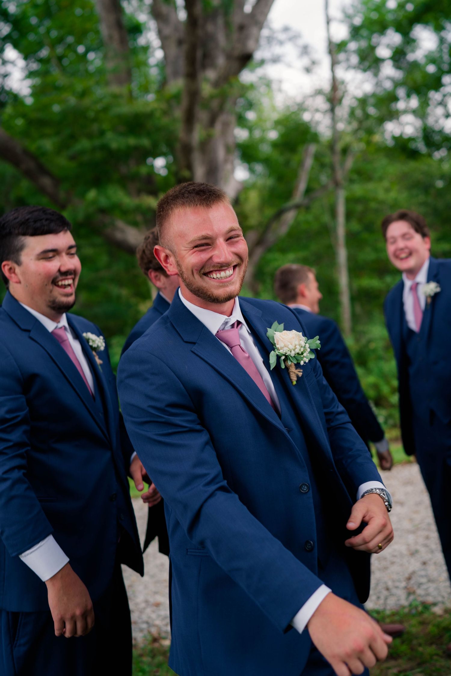 Smiling groom in navy blue suit with pink tie and boutonniere walks with laughing groomsmen at outdoor wedding.