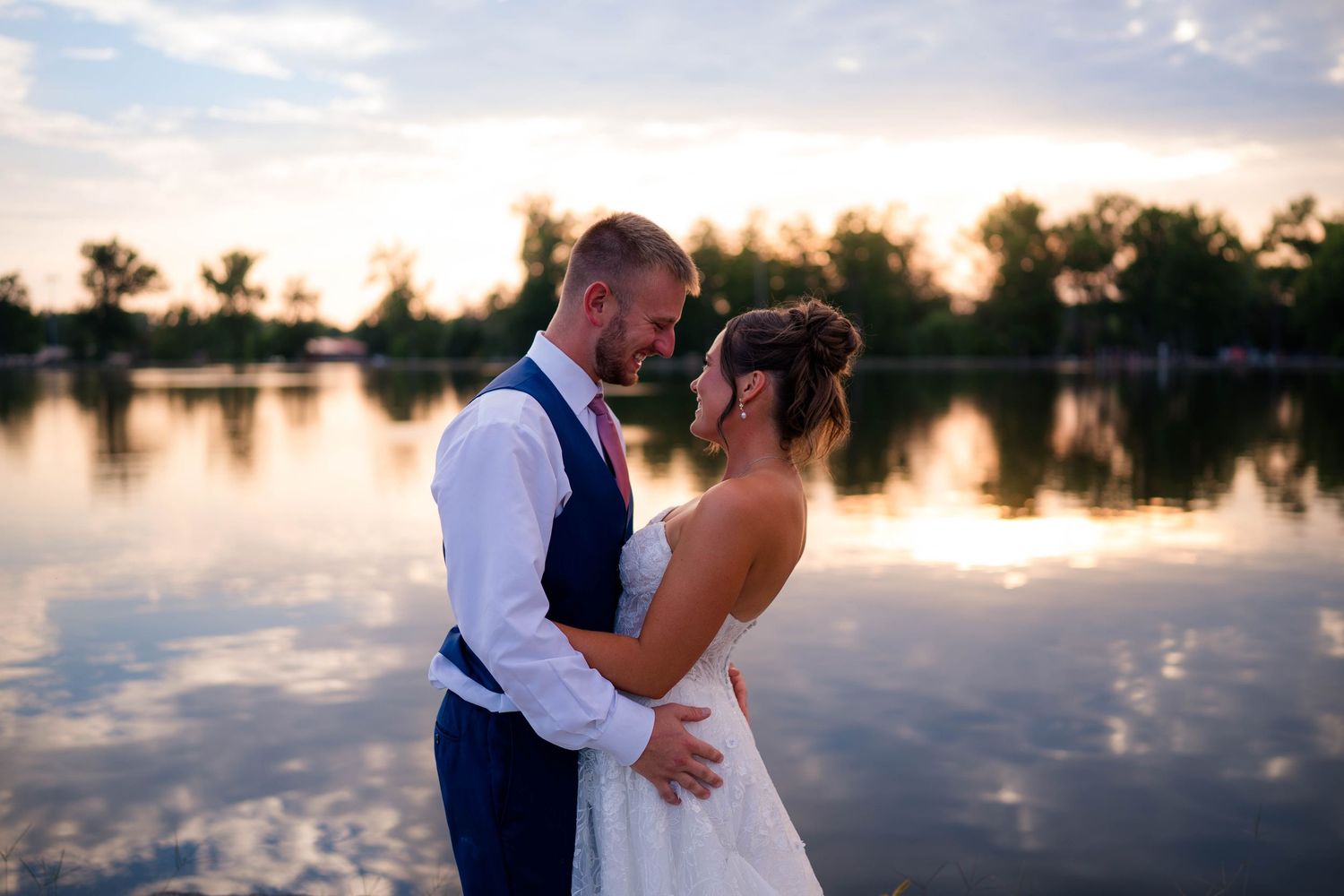 Couple embraces at sunset by a reflective lake, groom in vest and bride in white gown, trees silhouetted behind them.