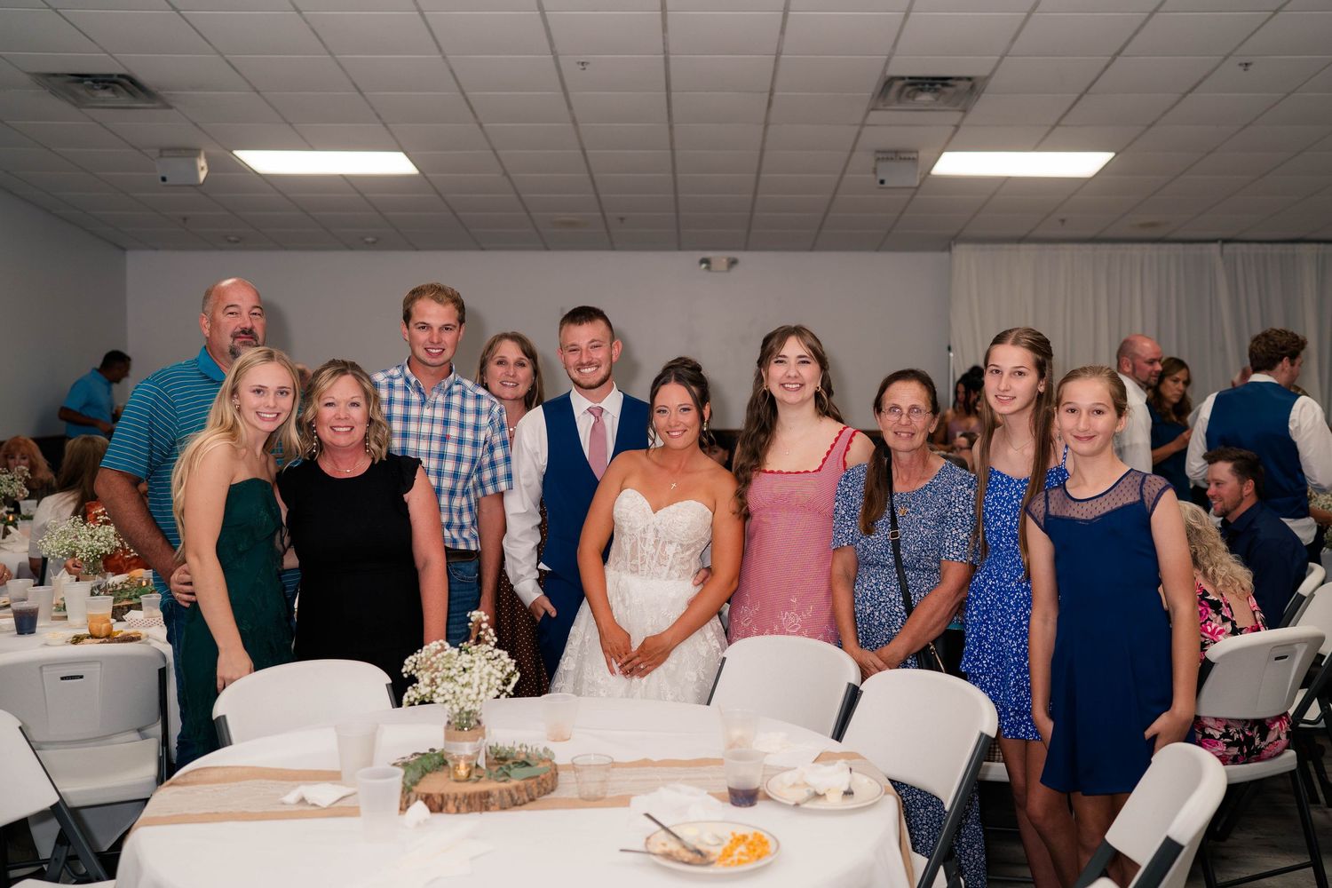 Wedding party and family members pose together smiling at a reception venue with white chairs and decorated tables.