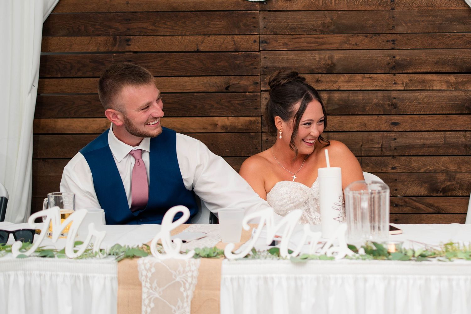 Bride and groom laugh together at sweetheart table decorated with Mr. & Mrs. sign and greenery at wedding reception.