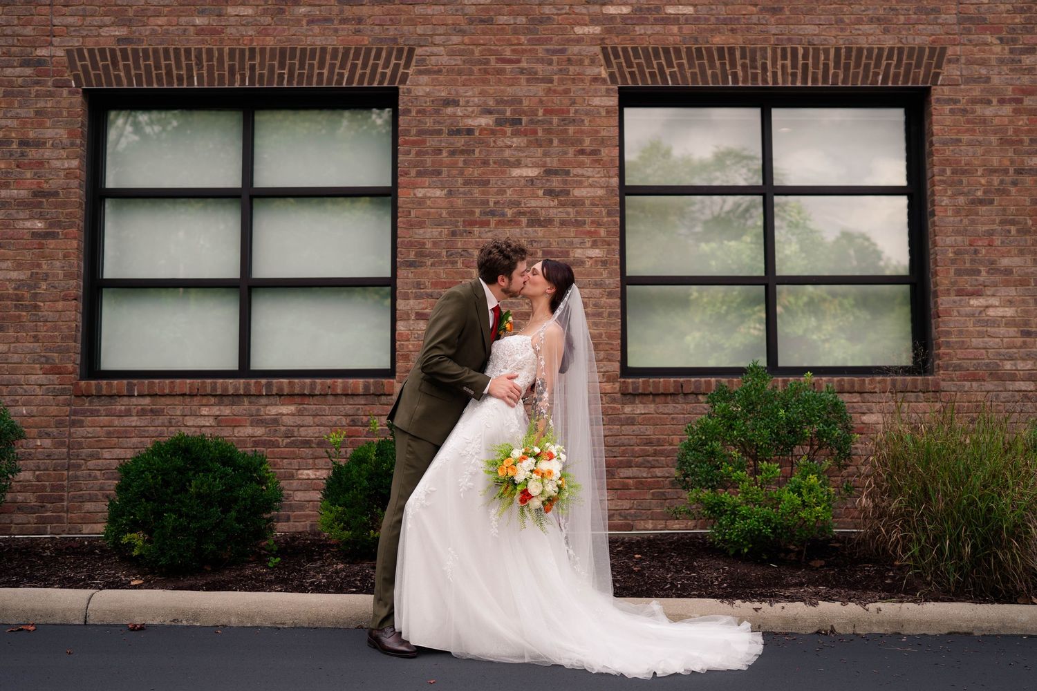 Bride and groom share a romantic kiss outside a brick building, bride holding a colorful orange and white bouquet.