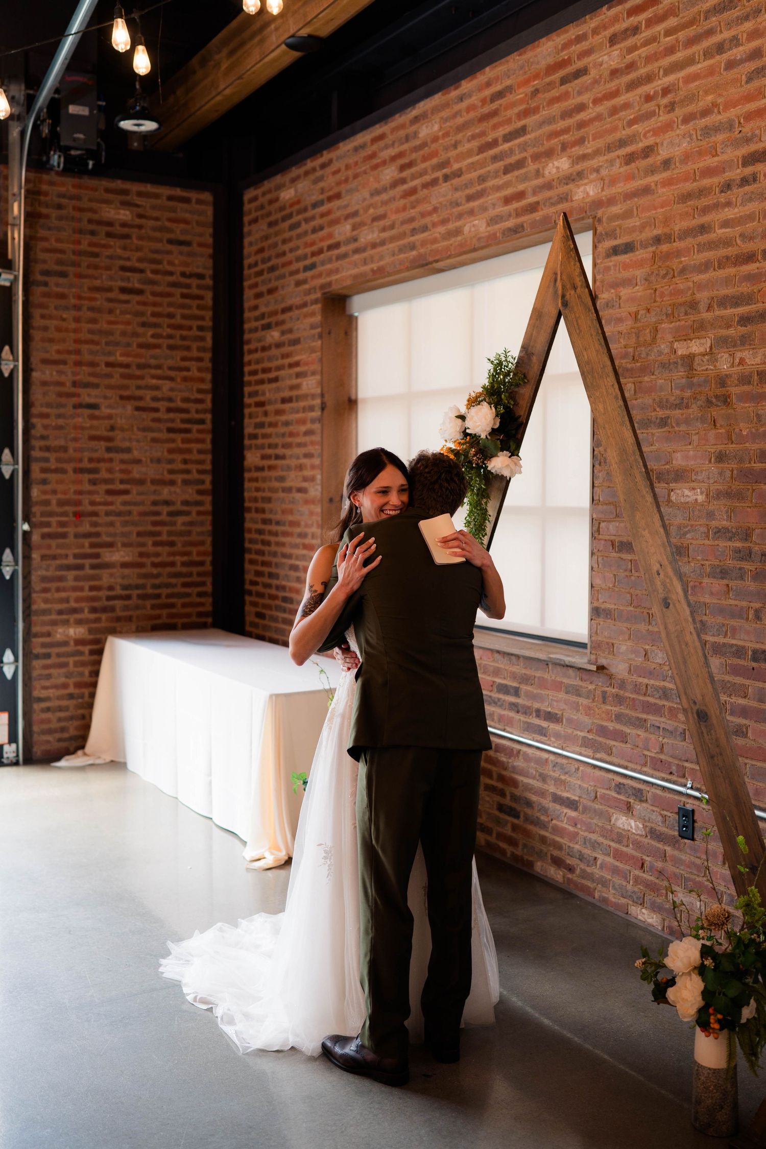 Bride and groom share a tender embrace inside a rustic brick venue with a wooden arch backdrop.
